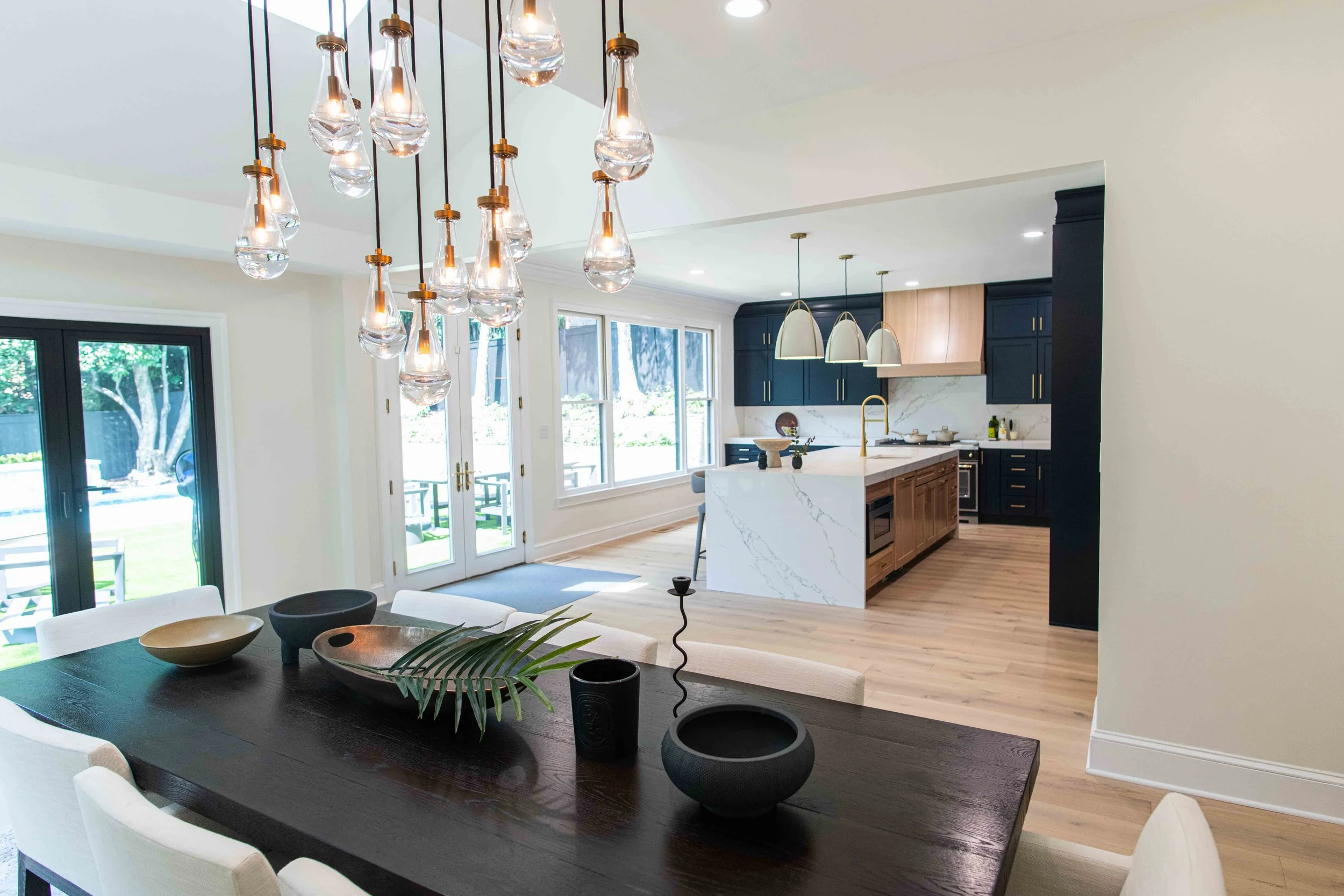 Modern open-concept kitchen and dining area with black and wooden cabinets, white marble countertops, a kitchen island, and a contemporary chandelier above a dark wood dining table decorated with bowls, a leaf, and a candle. Large windows and glass d