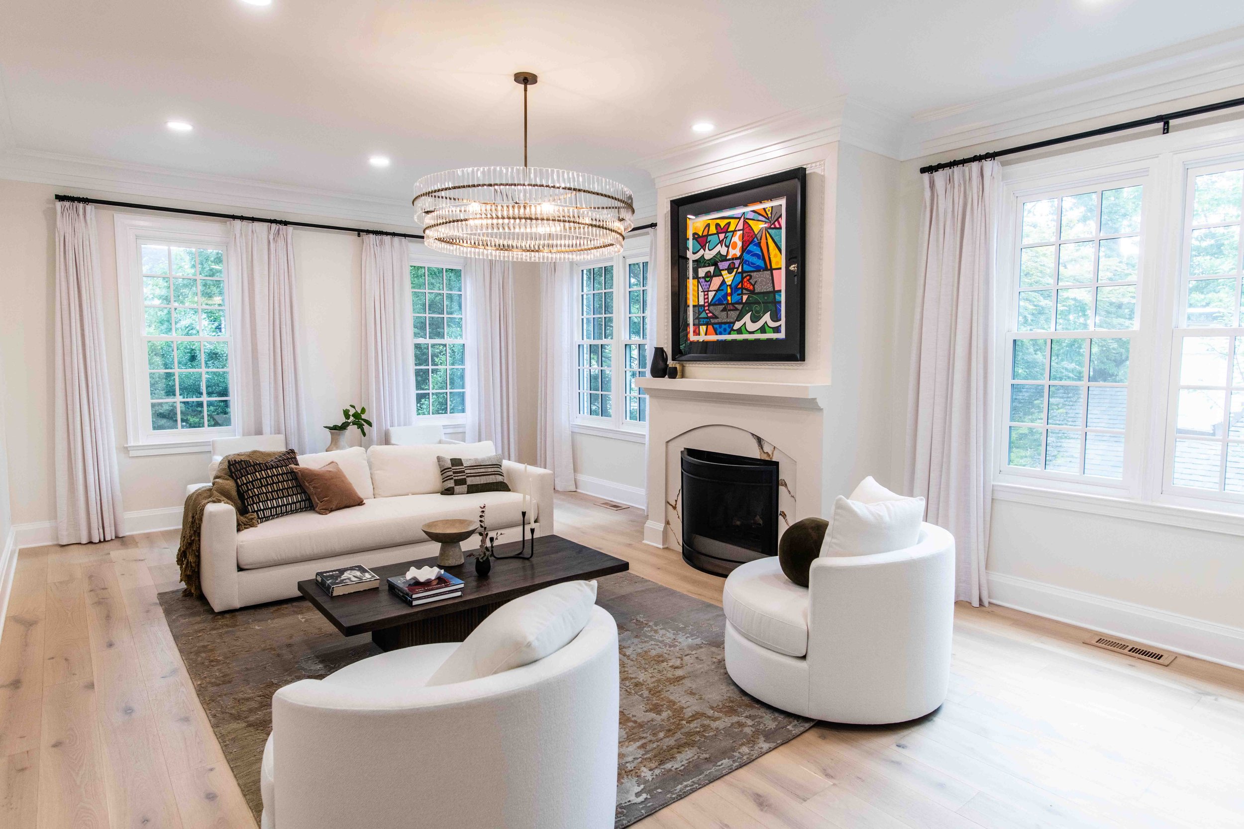 Bright living room with white walls, large windows with white curtains, a chandelier, and contemporary furniture including a white sofa, white armchairs, and a black coffee table. There is a colorful abstract painting above the fireplace.
