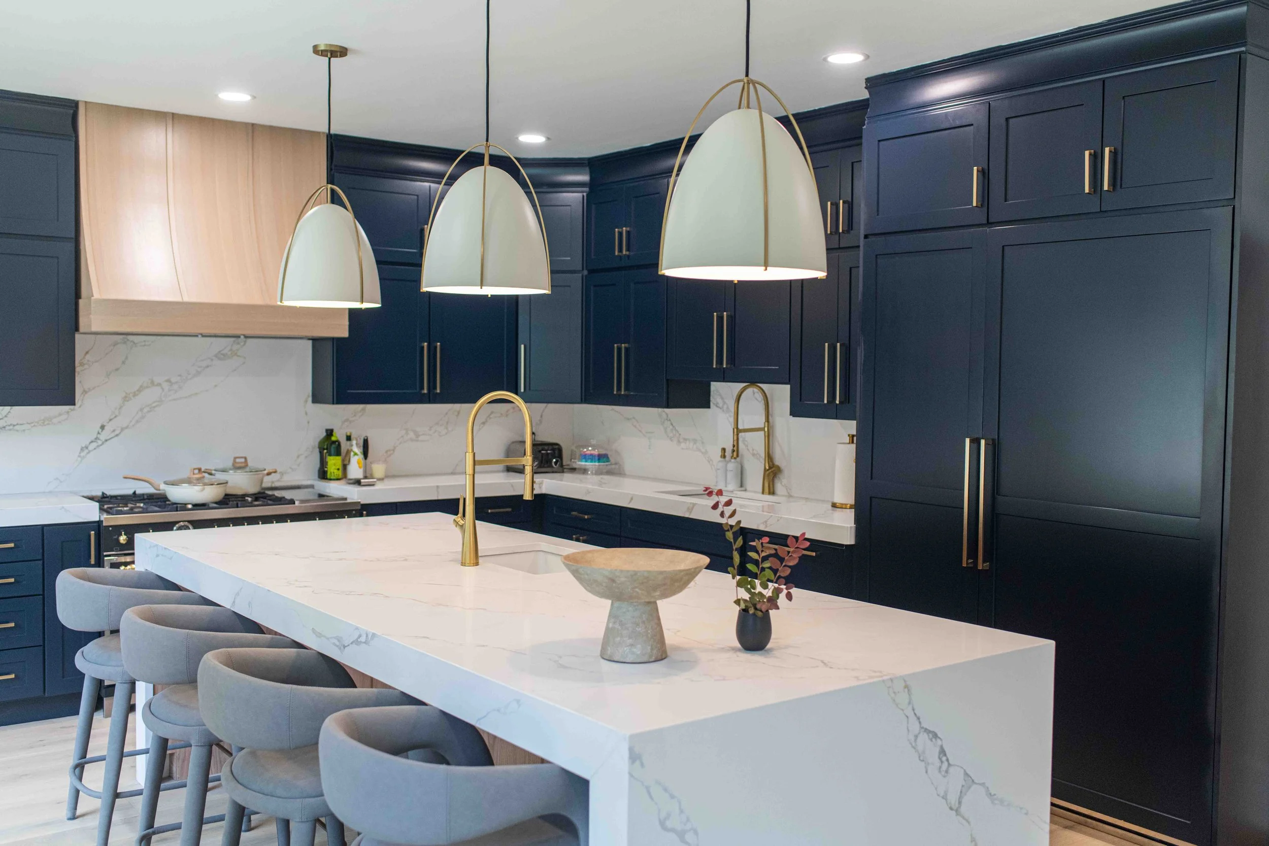 Modern kitchen with dark navy cabinets, white marble countertops, and a large central island with barstools. Features gold fixtures, pendant lights, and a gold faucet.