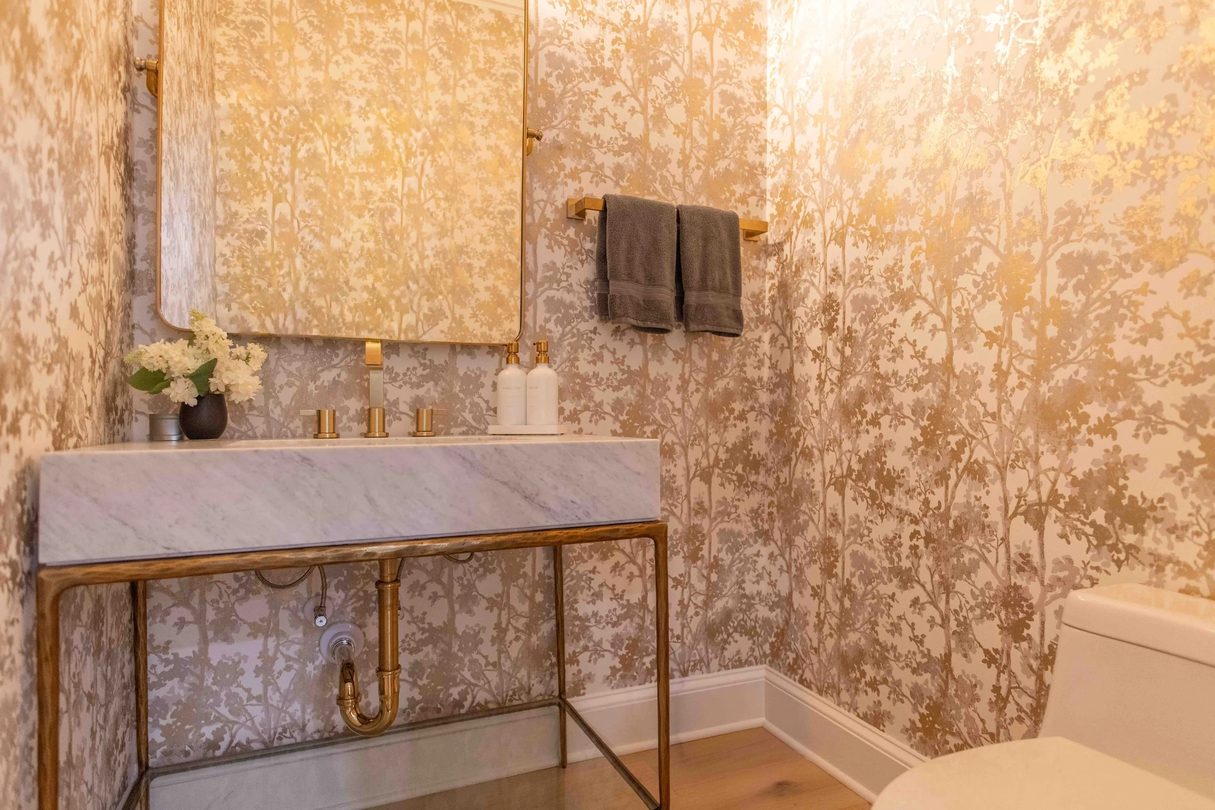 Bathroom with a marble sink, gold fixtures, a mirror, two gray towels on a gold towel rack, a small vase with white flowers, and wallpaper with a gold tree pattern.