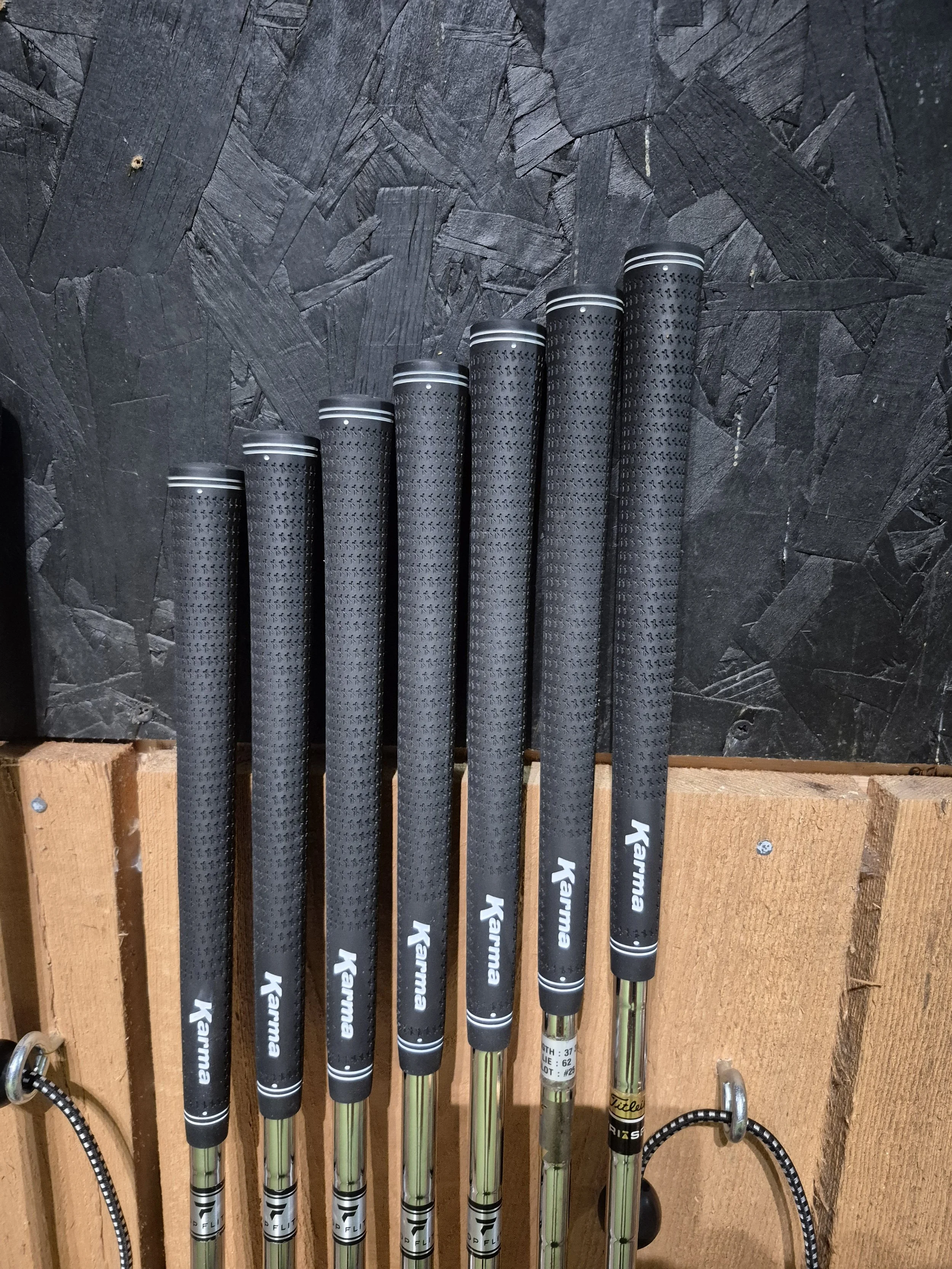 Six golf clubs with black grips and chrome shafts hanging on a wooden wall against a dark stone background.