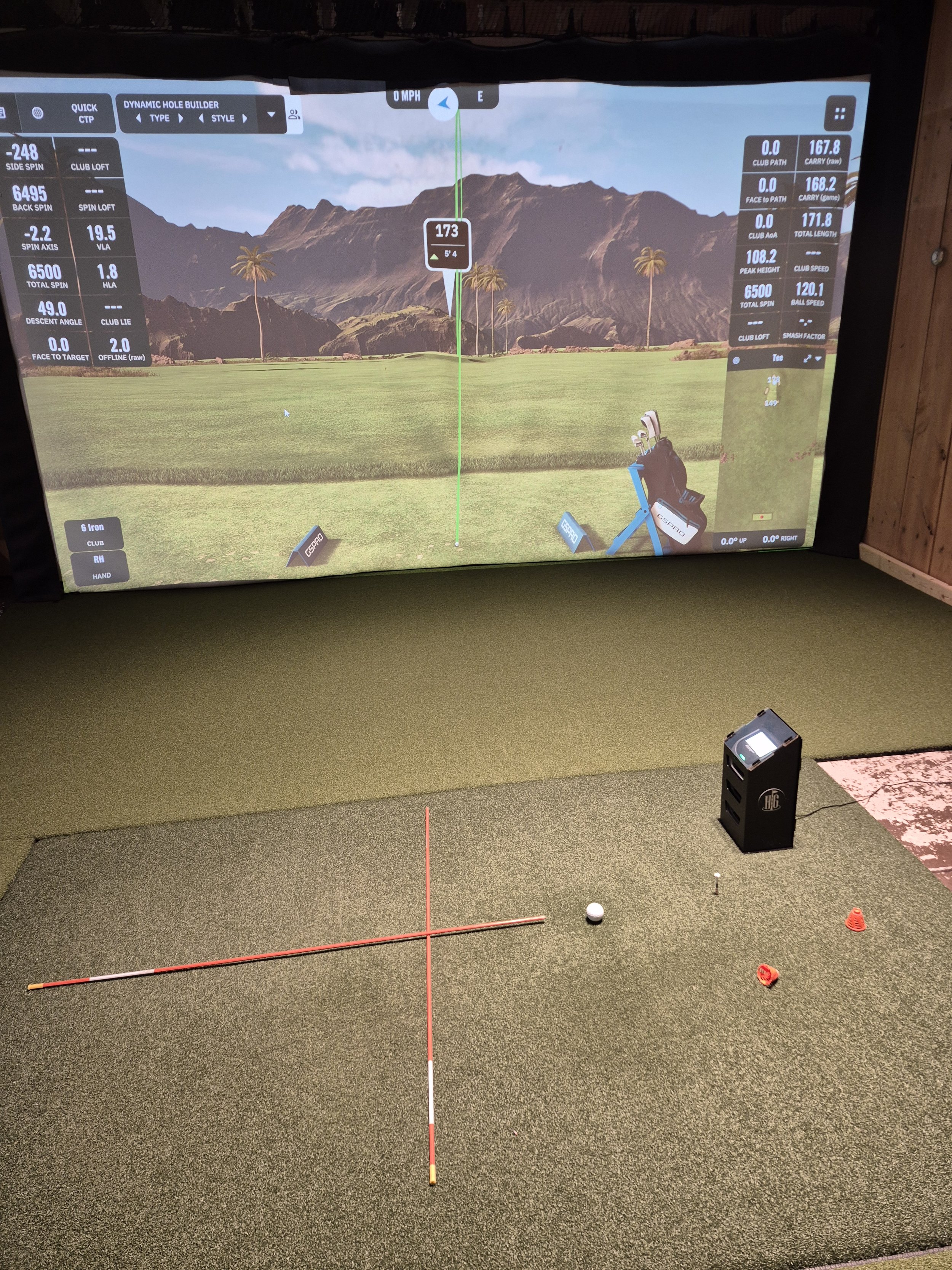 Indoor golf simulator with a screen showing a virtual golf course, a golf ball on the mat, and a golf club brace. The simulator displays data and analysis of a golf swing.