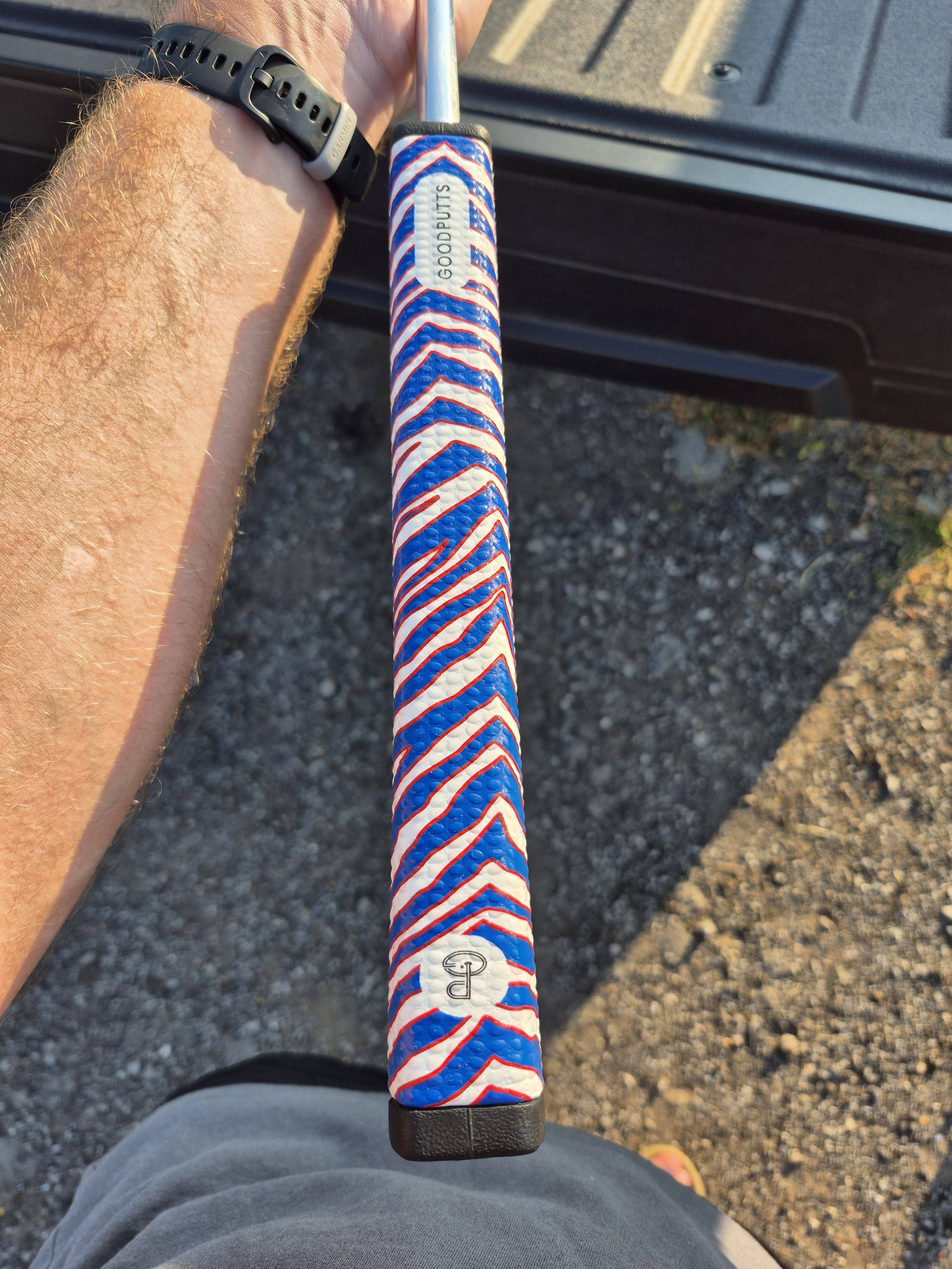 A golf club with a red, white, and blue striped grip and a label that says 'Good Putts,' held by a person wearing a black wristwatch, with a gravel surface and part of a black vehicle in the background.
