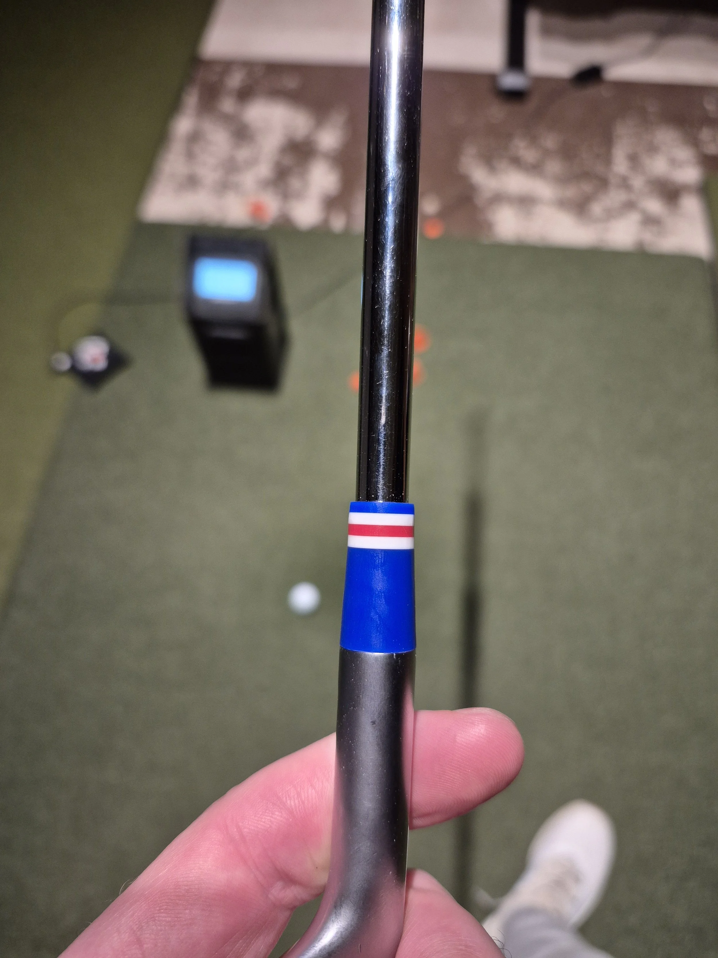 Close-up of a golf club held by a person, with a golf ball and golf course equipment visible in the background.