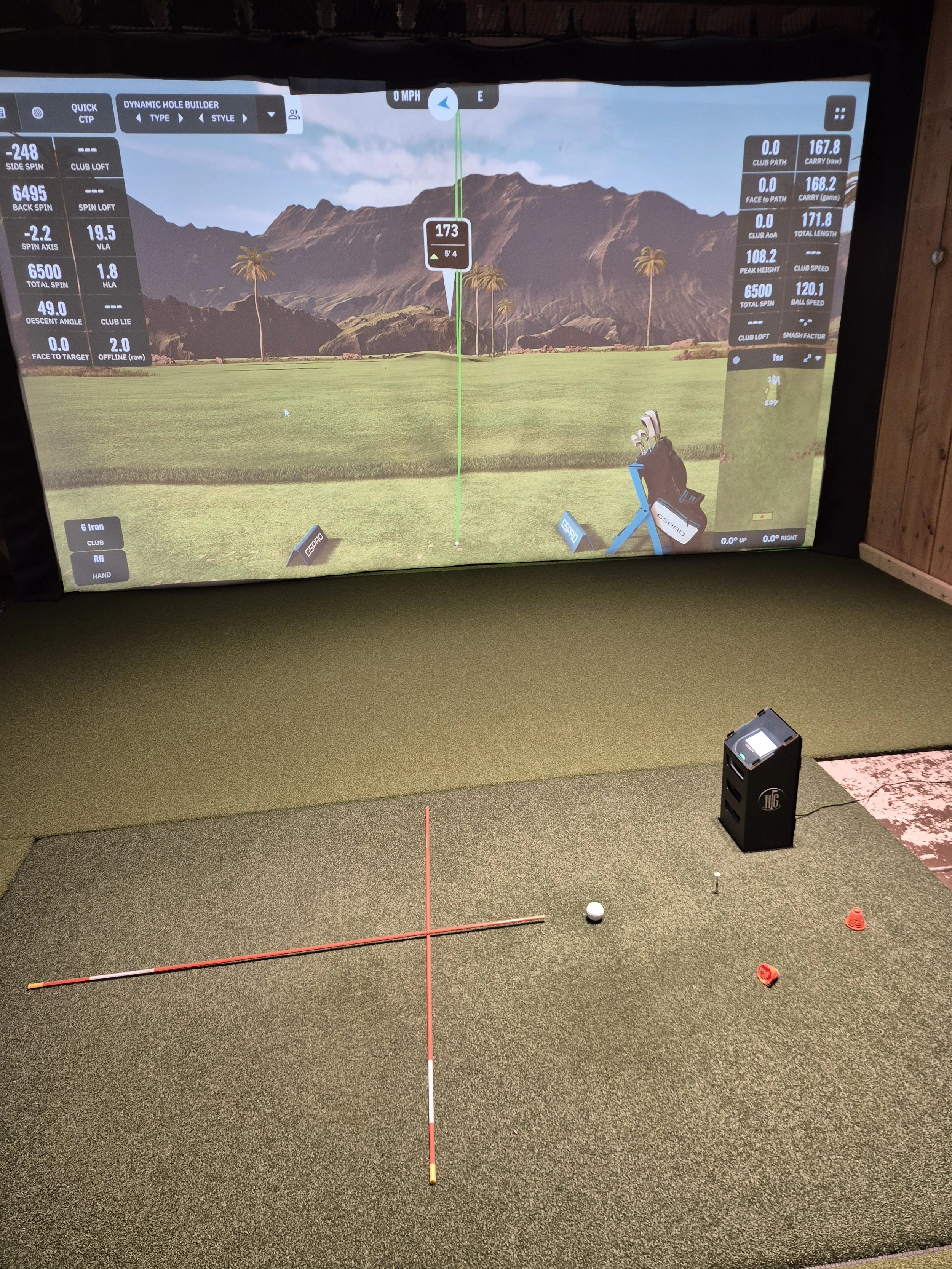Golf simulator setup with a golf ball, alignment rods, a hitting mat, and a large screen displaying a virtual golf course landscape.
