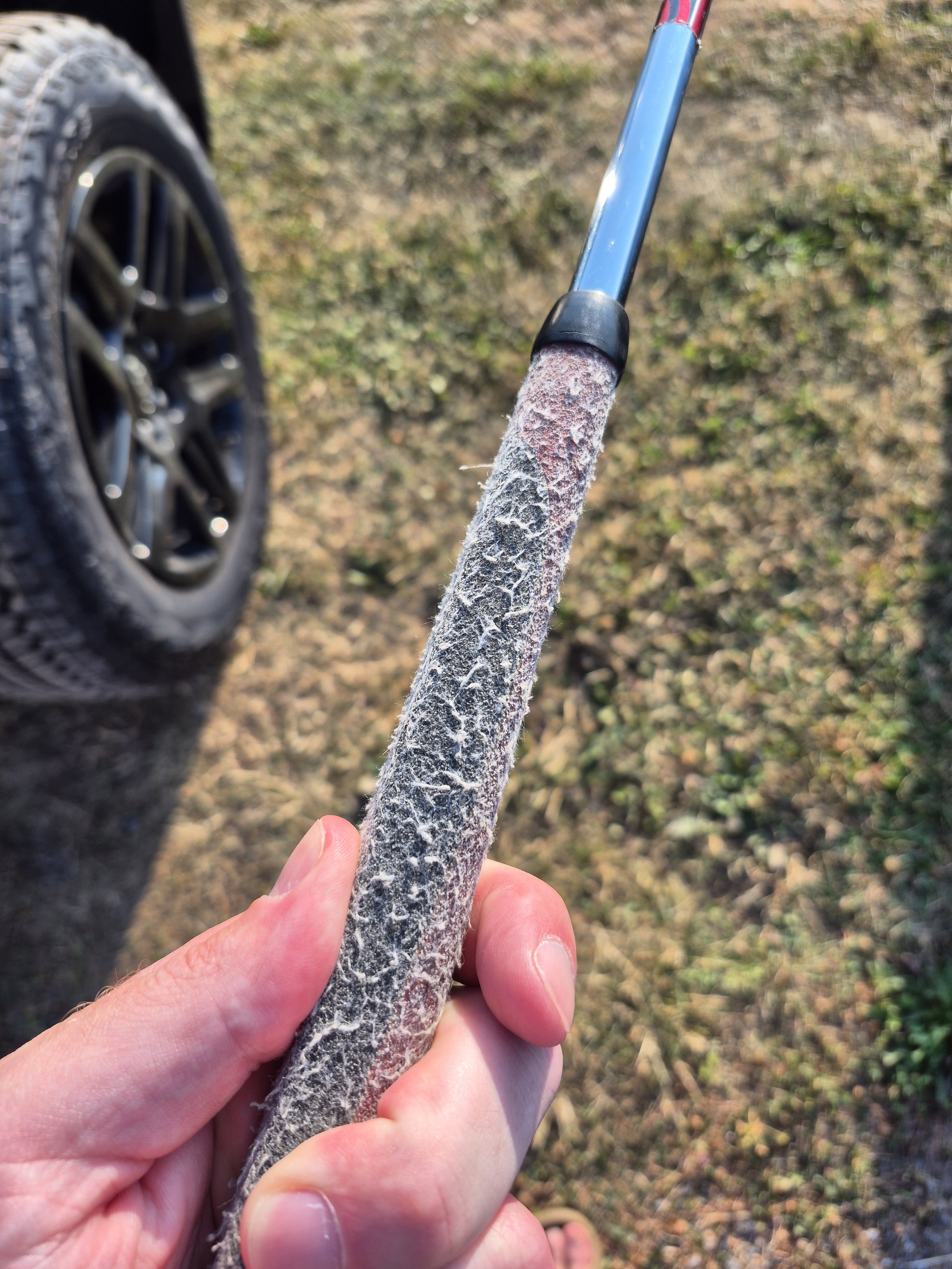Close-up of a hand holding a golf club covered in frost, with a tire and grassy ground in the background.