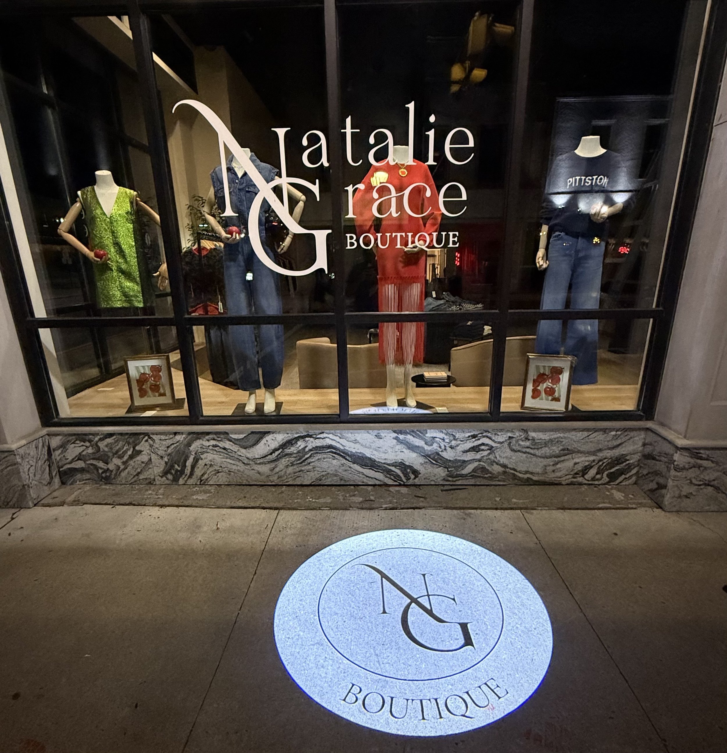 Storefront window display for Natalie Grace Boutique featuring mannequins dressed in colorful and denim clothing, with the boutique's logo and name visible on the glass and a circular light projection on the sidewalk.