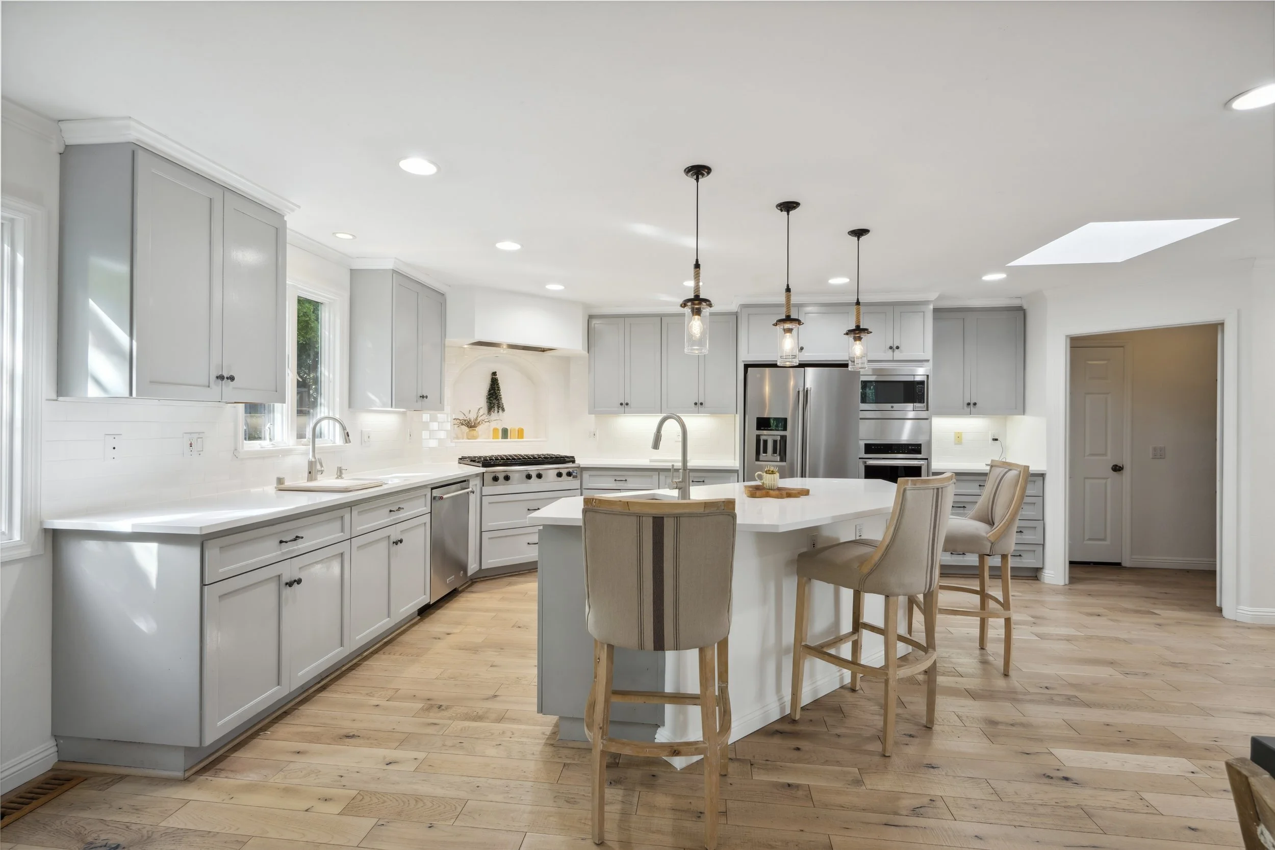 Modern white kitchen with gray cabinets, a kitchen island with bar stools, stainless steel appliances, hardwood floors, and pendant lights hanging above the island.