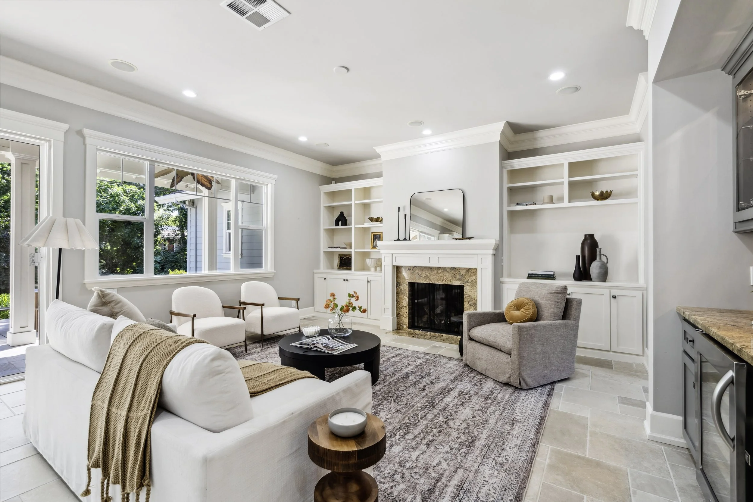 Bright living room with white walls, a white sofa with beige throw, two white armchairs, and a gray armchair around a black round coffee table. Large window with trees outside, built-in white bookshelves, and a fireplace with a mirror above. Light-colored tiled floor and a patterned area rug.