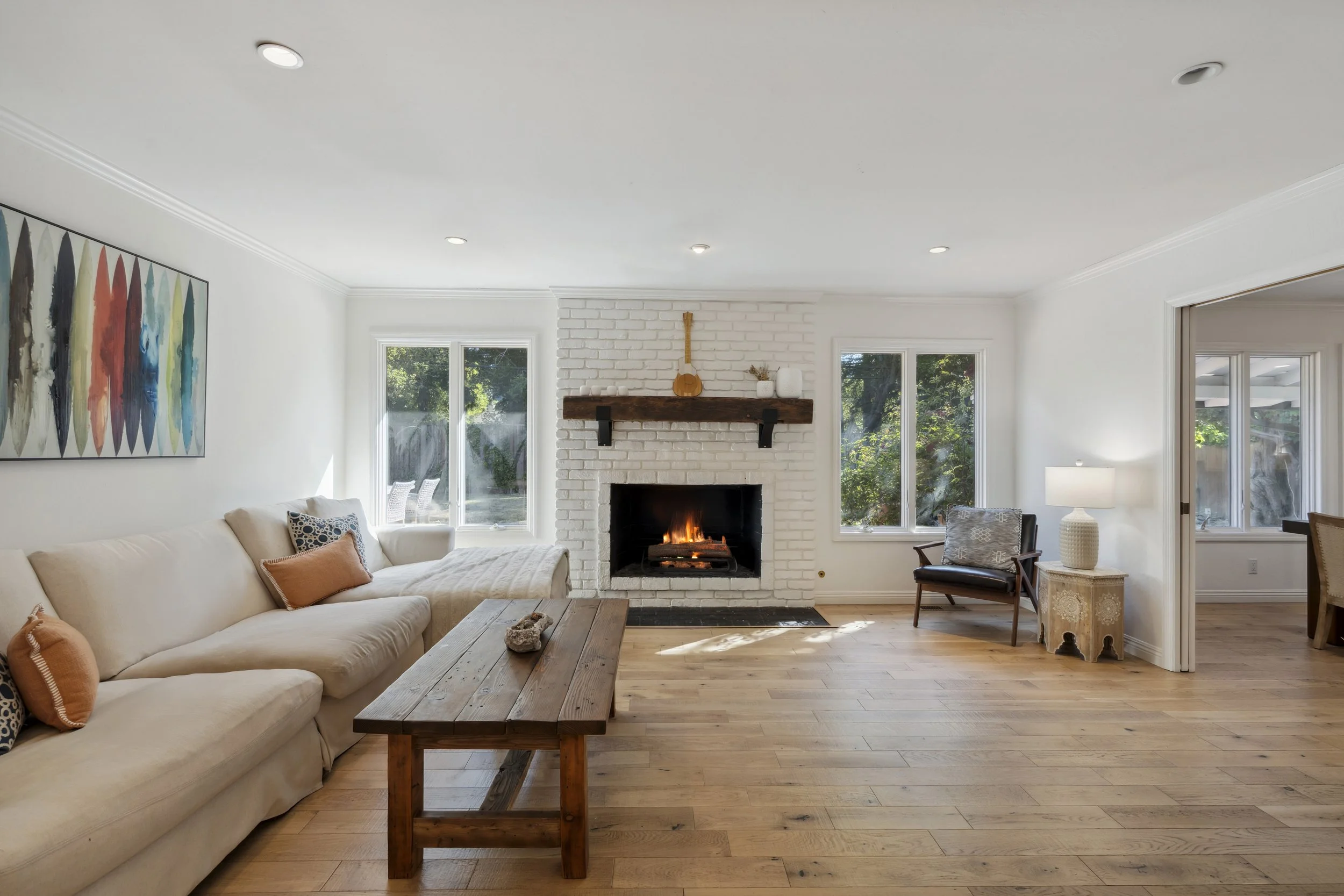 Living room with a white brick fireplace, wooden mantel, beige sectional sofa with throw pillows, wooden coffee table, armchair with patterned cushion, side table with lamp, and large windows showing outdoor greenery.