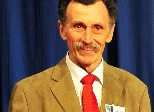 Homme souriant avec une moustache, portant un costume beige, une chemise blanche et une cravate rouge, posant devant un rideau bleu.