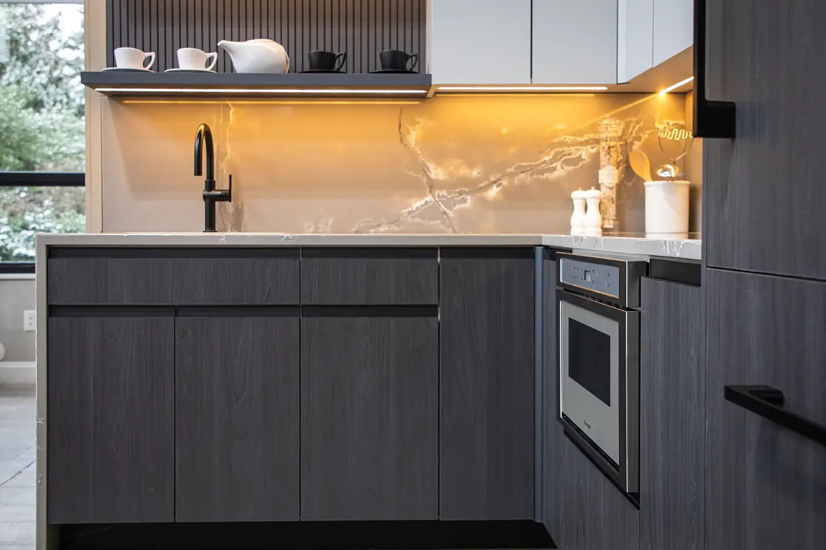 Modern kitchen with dark wood cabinets, marble backsplash, and a white countertop featuring a black faucet. Open shelves with white and black mugs and a teapot. Built-in oven and decorative accents on the counter.