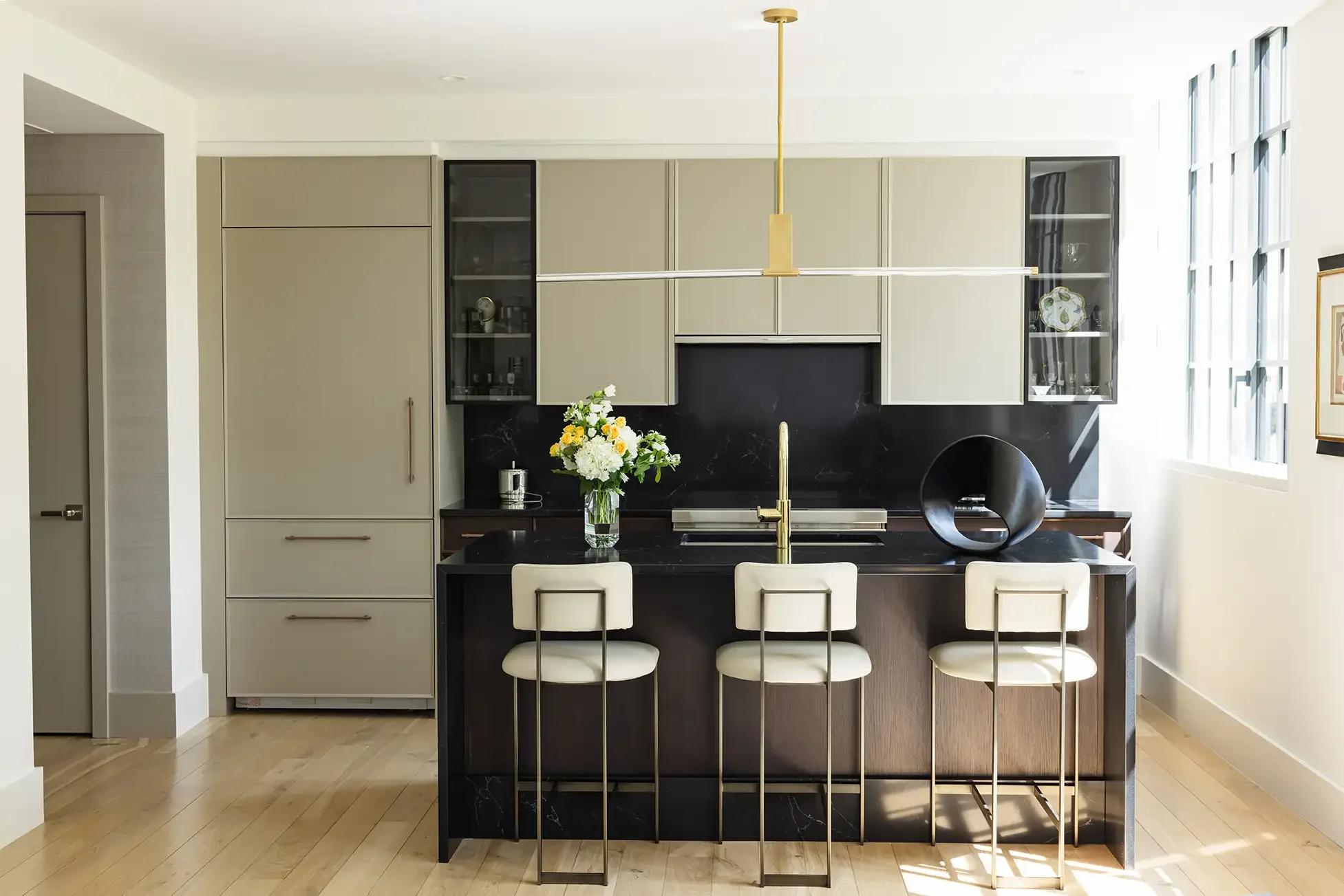 Modern kitchen with beige cabinetry, black countertops, a black backsplash, and a kitchen island with four barstools, a vase of flowers, and decorative items. Sunlight streams through large windows on the right.