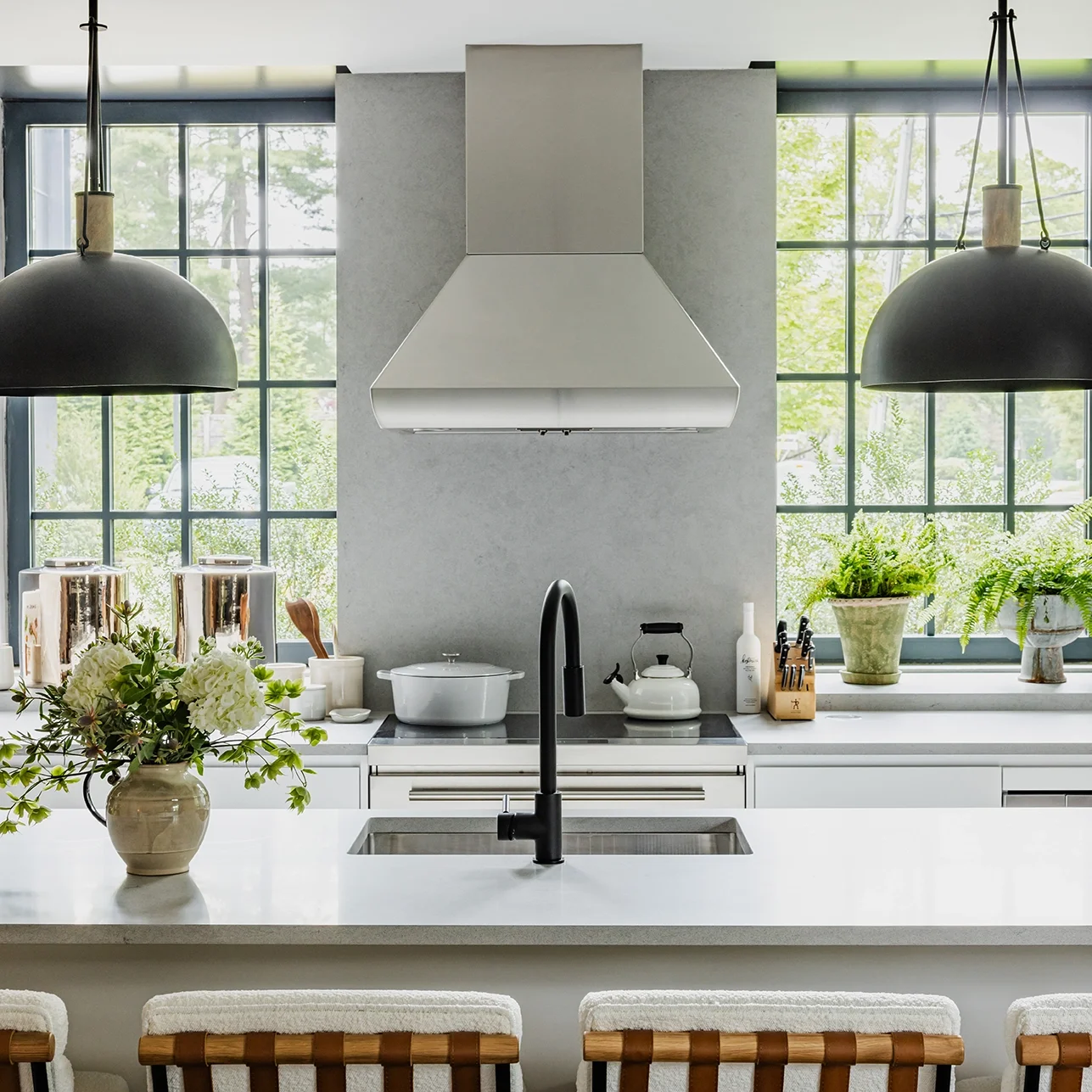 Modern kitchen with white countertops, black faucet, and large windows with greenery outside. Decor includes potted plants, flowers, and kitchen utensils.