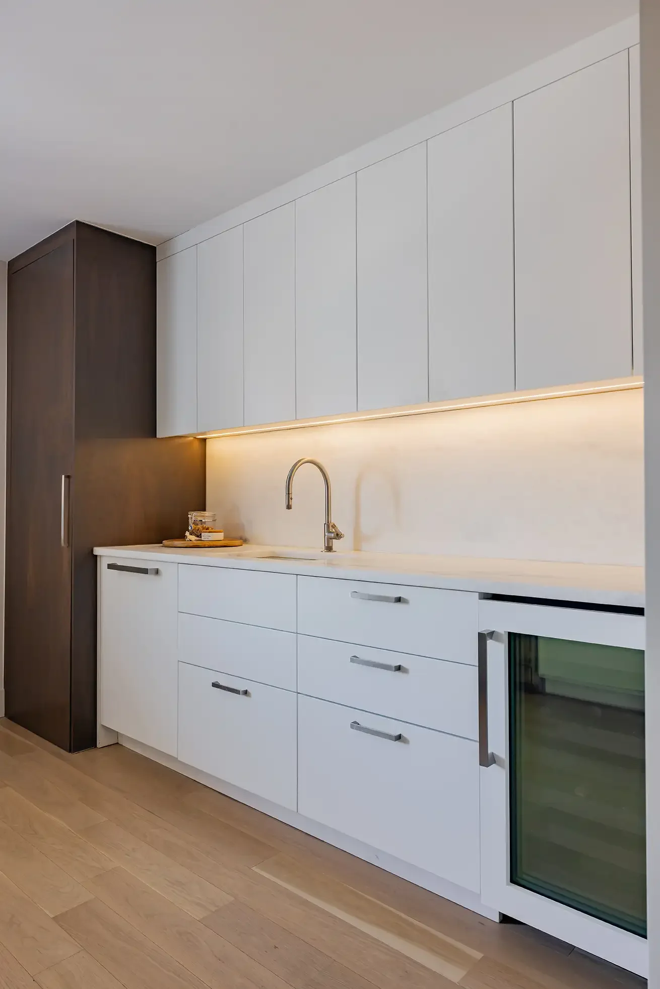 Modern kitchen with white cabinets, a built-in fridge, and a small wine cooler on the right. The countertop has a silver faucets sink, and there is a small jar and tray with cookies on the corner. The upper cabinets are white, and the back wall has a