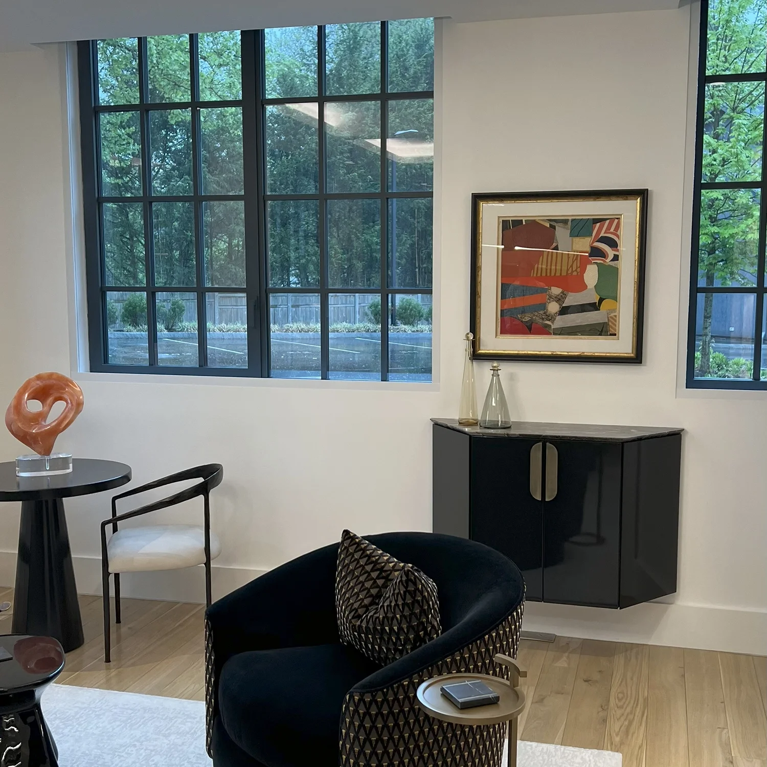 Modern living room with large black-framed windows, a decorated black cabinet, abstract artwork on the wall, and seating including a black velvet armchair with a patterned pillow and a white and black chair near a round table with a decorative sculpt