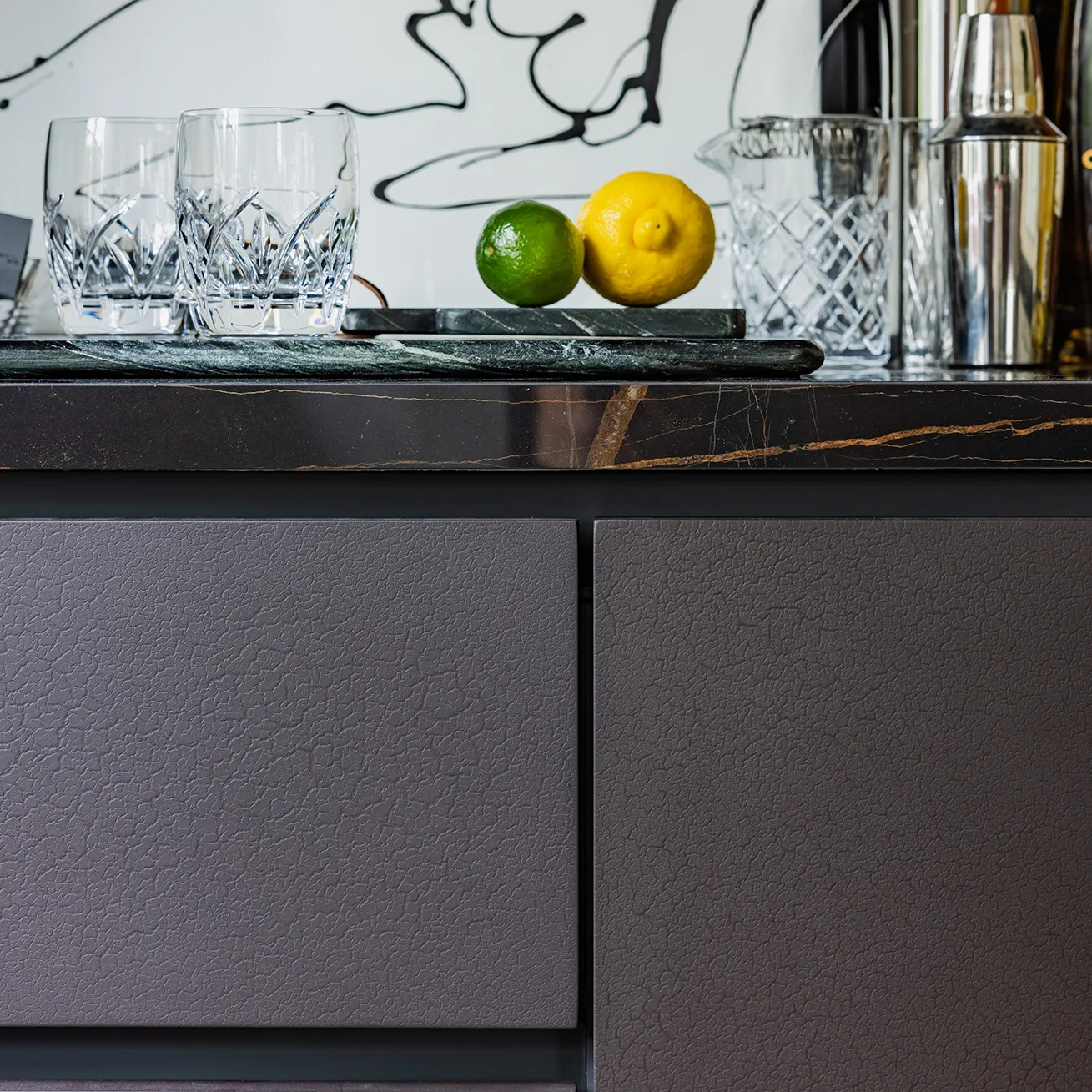 A modern kitchen counter with two glasses of water, a lime, a lemon on a tray, and a glass pitcher, with a black and white abstract art in the background.
