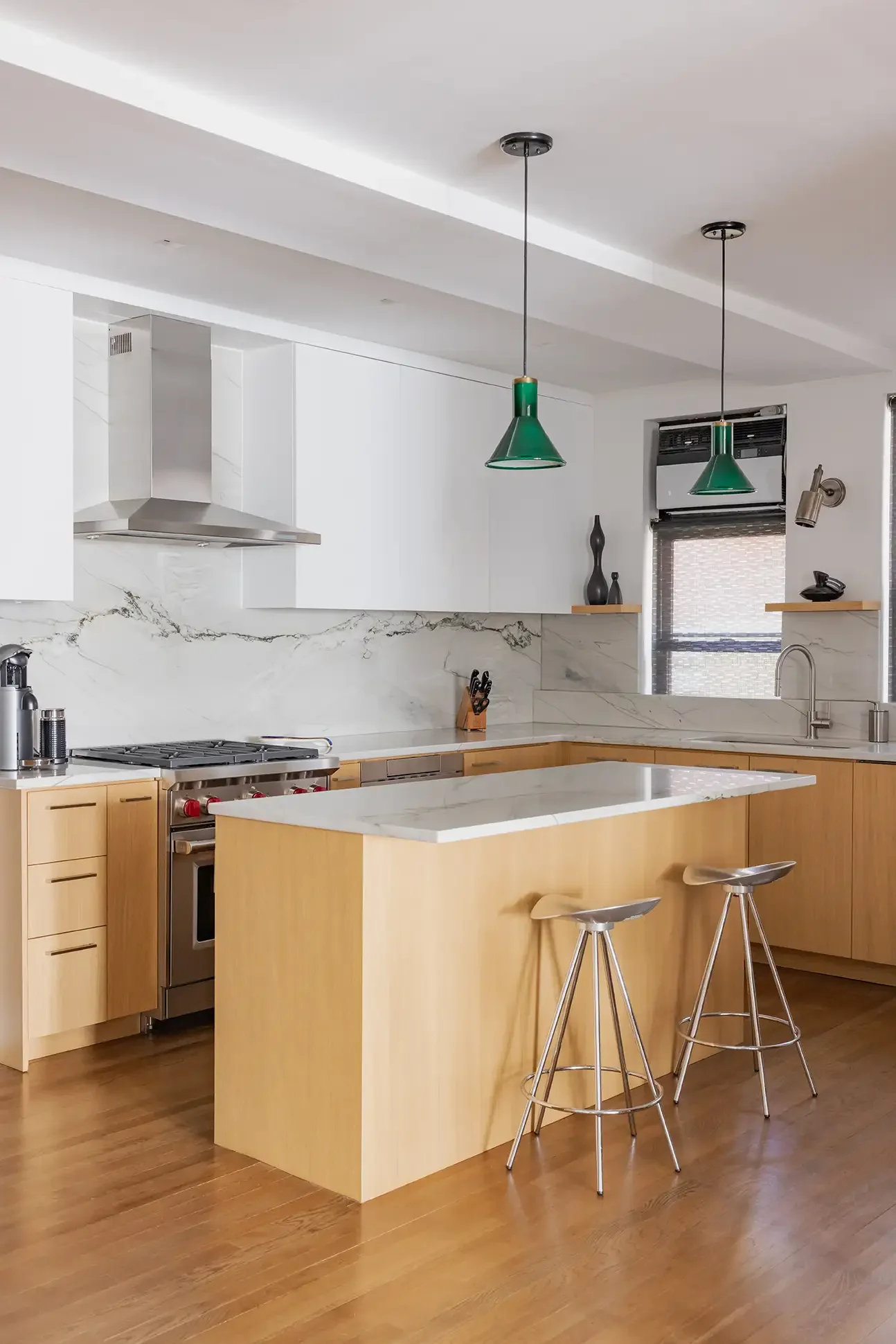 Modern kitchen with white cabinets, marble backsplash, wooden island with marble top, two green pendant lights, and gray bar stools.