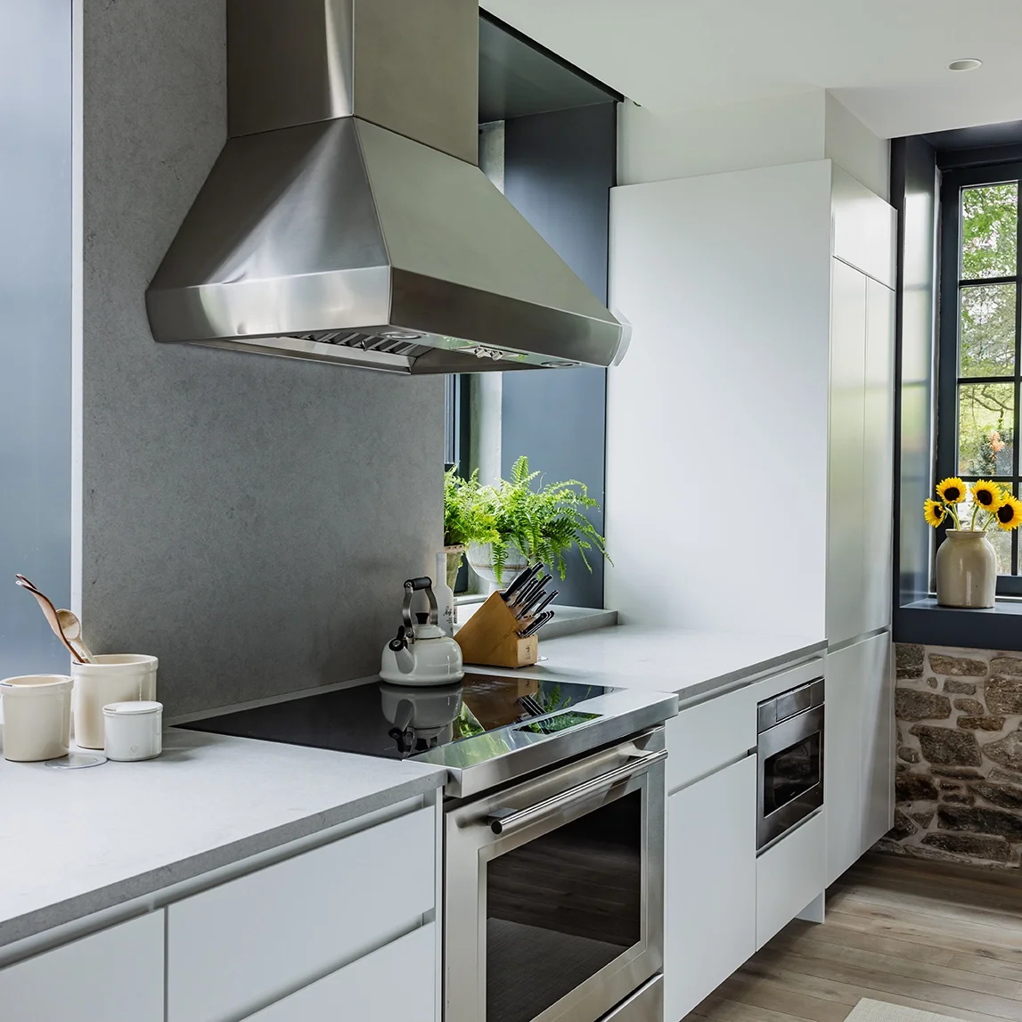 Modern kitchen with stainless steel range hood, white cabinetry, black cooktop, potted fern, and a window with a sunflower vase.
