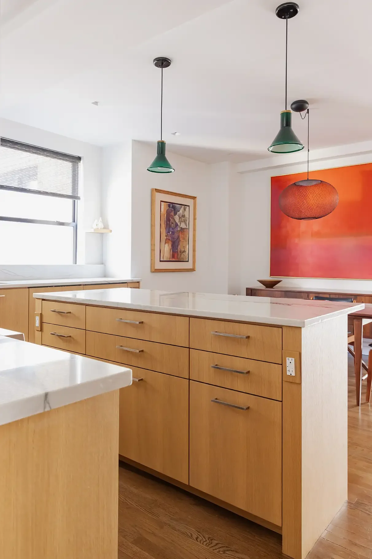 Modern kitchen with wooden cabinets, a white marble countertop, colorful hanging pendant lights, an abstract painting on the wall, and a window providing natural light.