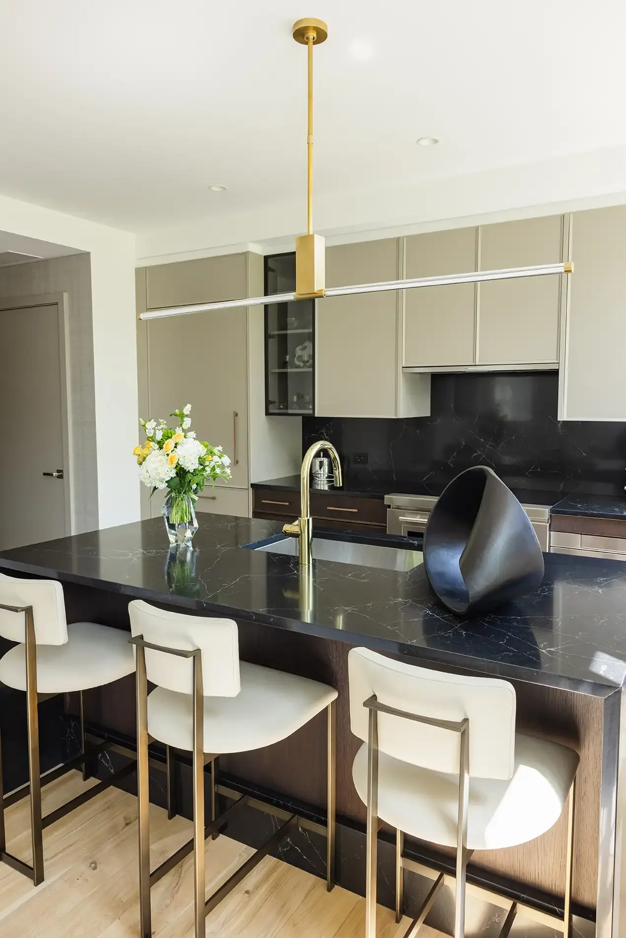 Modern kitchen with a black marble island, gold fixtures, white bar stools, a vase with white and yellow flowers, and a black decorative object.
