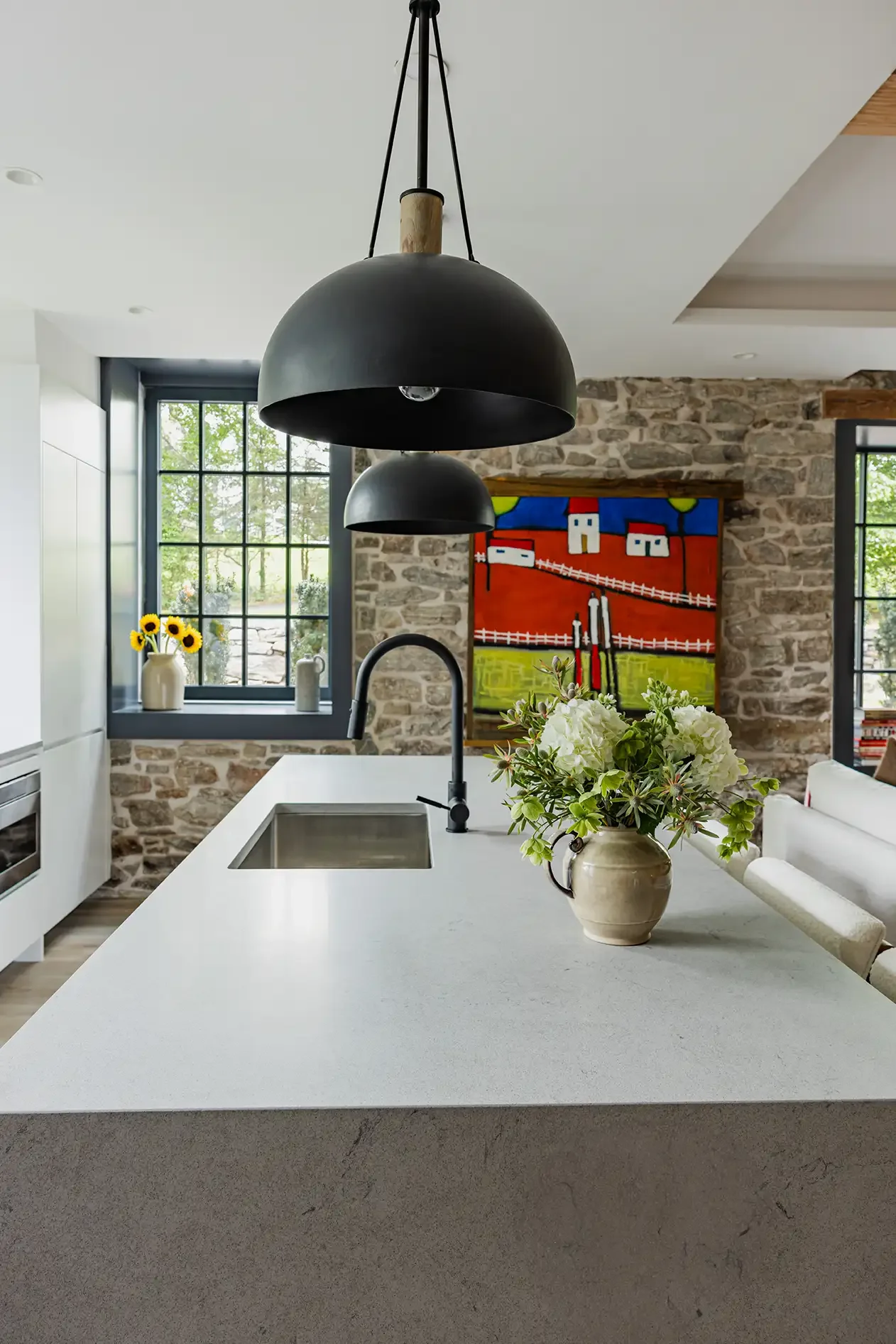 Modern kitchen with white countertop, black pendant lights, stone wall, colorful artwork, and a vase of flowers.