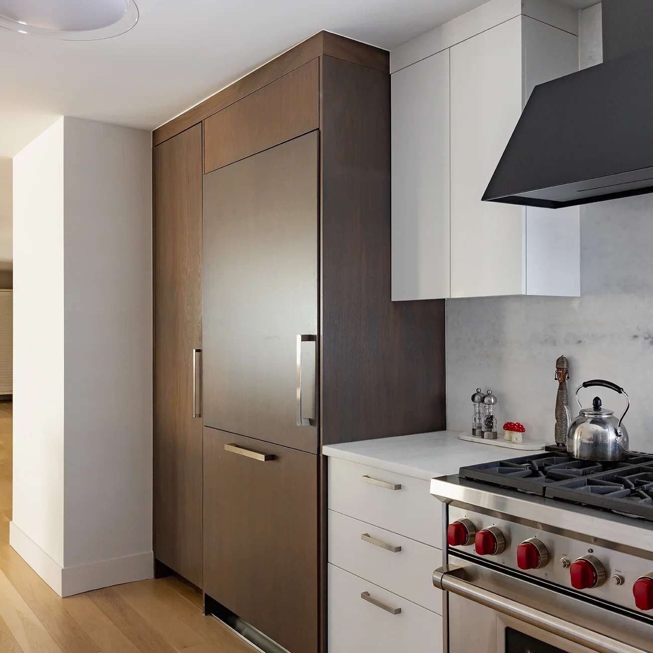 Kitchen with tall wooden refrigerator, white drawers, a stainless steel stove with red knobs, and a black range hood on a gray wall.