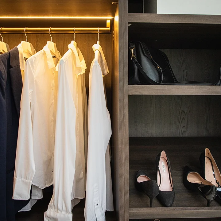 White shirts hanging in closet next to a shelf with black handbag and black high heels.
