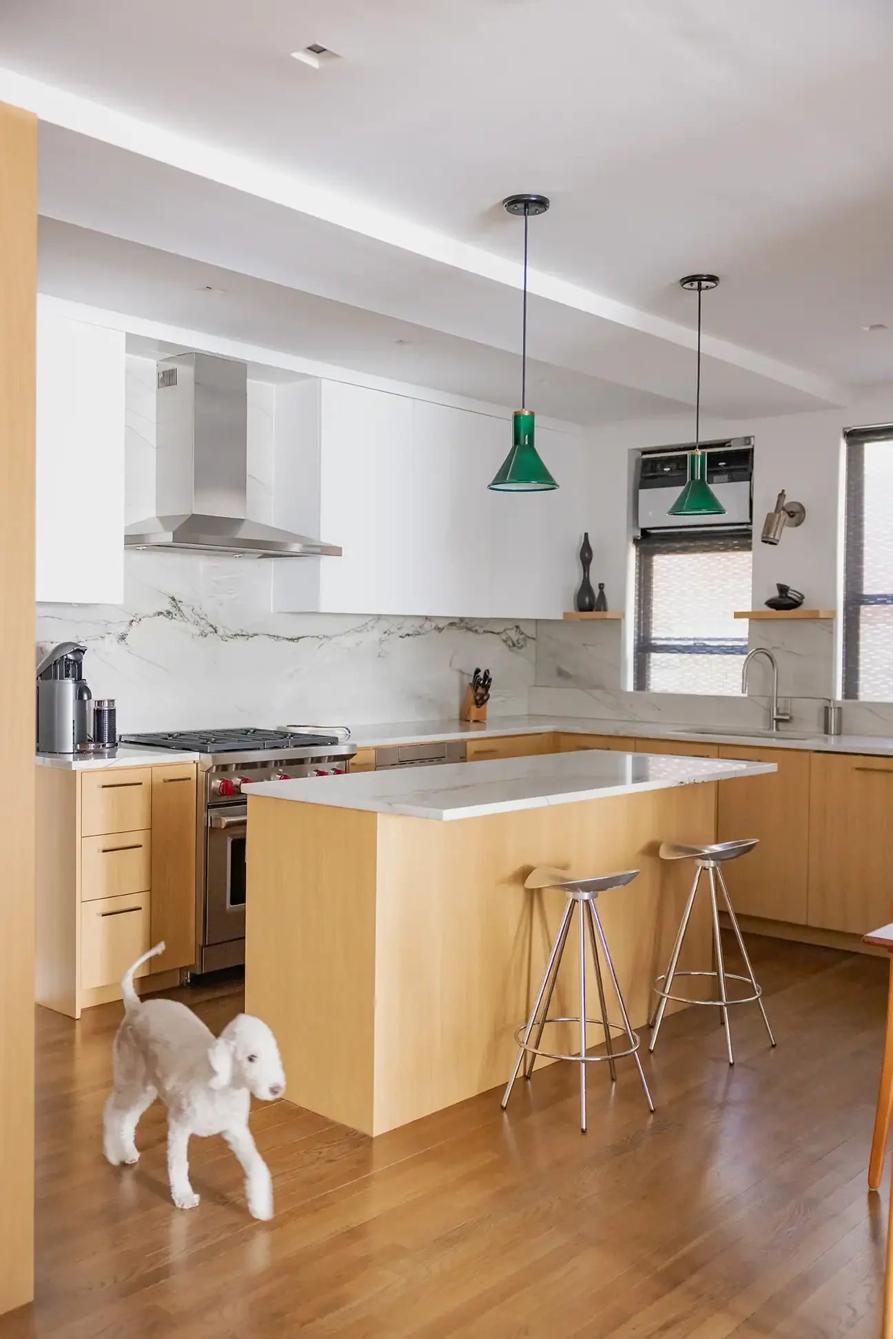 Modern kitchen with light wood cabinets, white marble countertops, and a white marble backsplash. Two green pendant lights hang above a kitchen island with a white marble top. There are two metal barstools at the island. A small white puppy is walkin