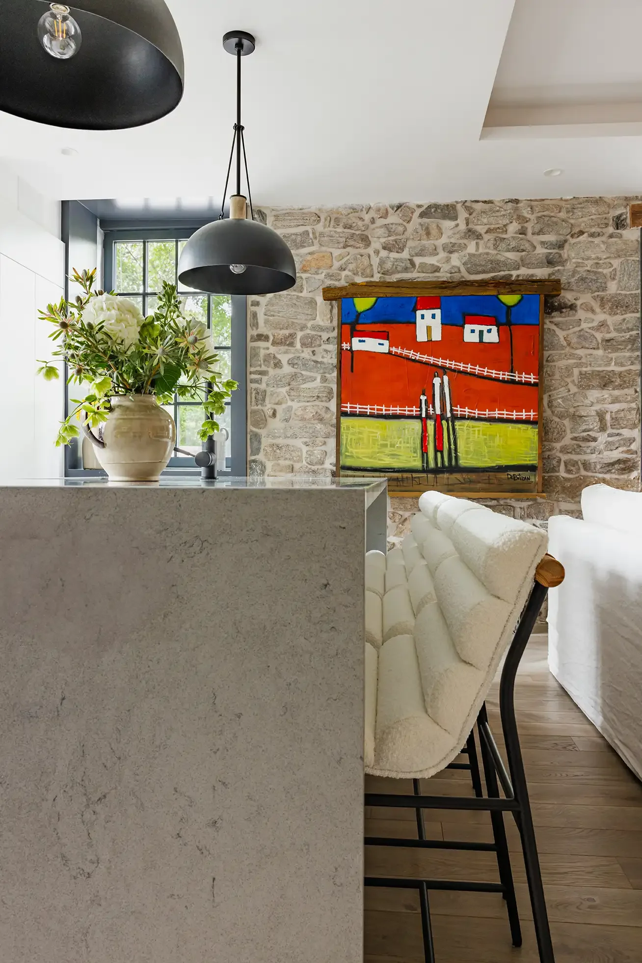 A modern kitchen with a beige stone wall, a black hanging pendant light, a window with black trim, a white counter with a beige vase of white flowers, and a colorful abstract painting on the stone wall. There are white upholstered bar stools with bla