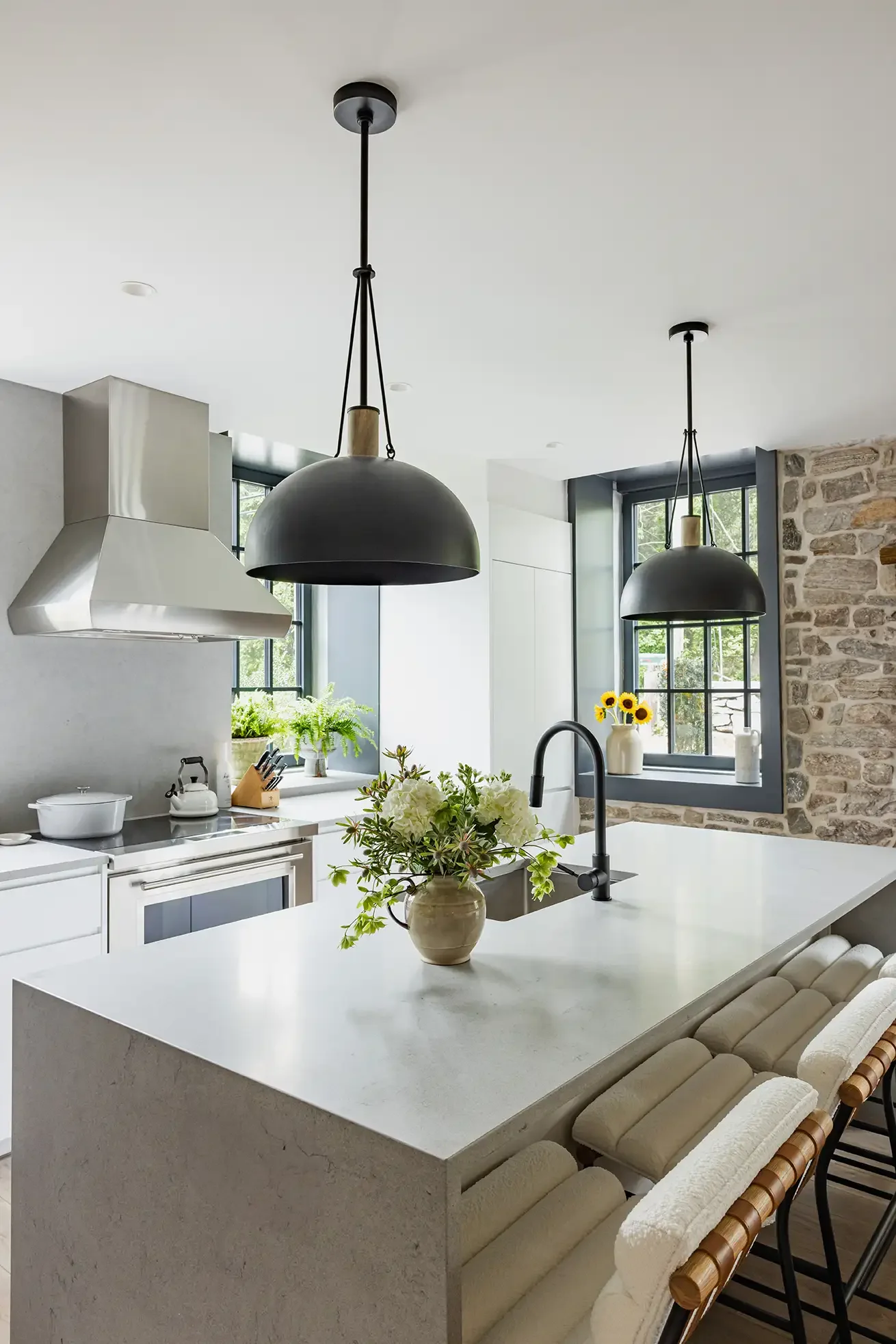Modern kitchen with white countertops, black pendant lights, a vase of flowers, a window with sunflowers, and a stone wall.