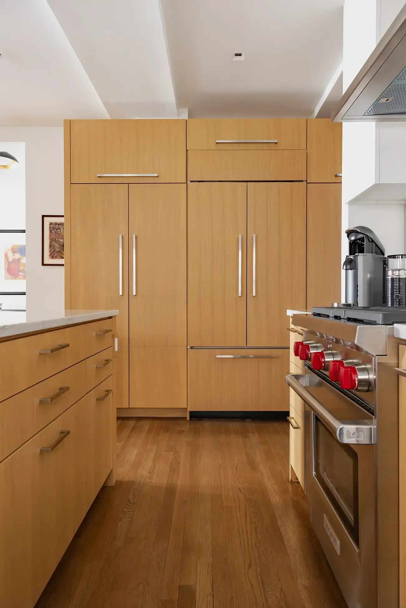 Modern kitchen with wooden cabinets, hardwood floors, a stainless steel stove with red knobs, and a coffee machine on the countertop.