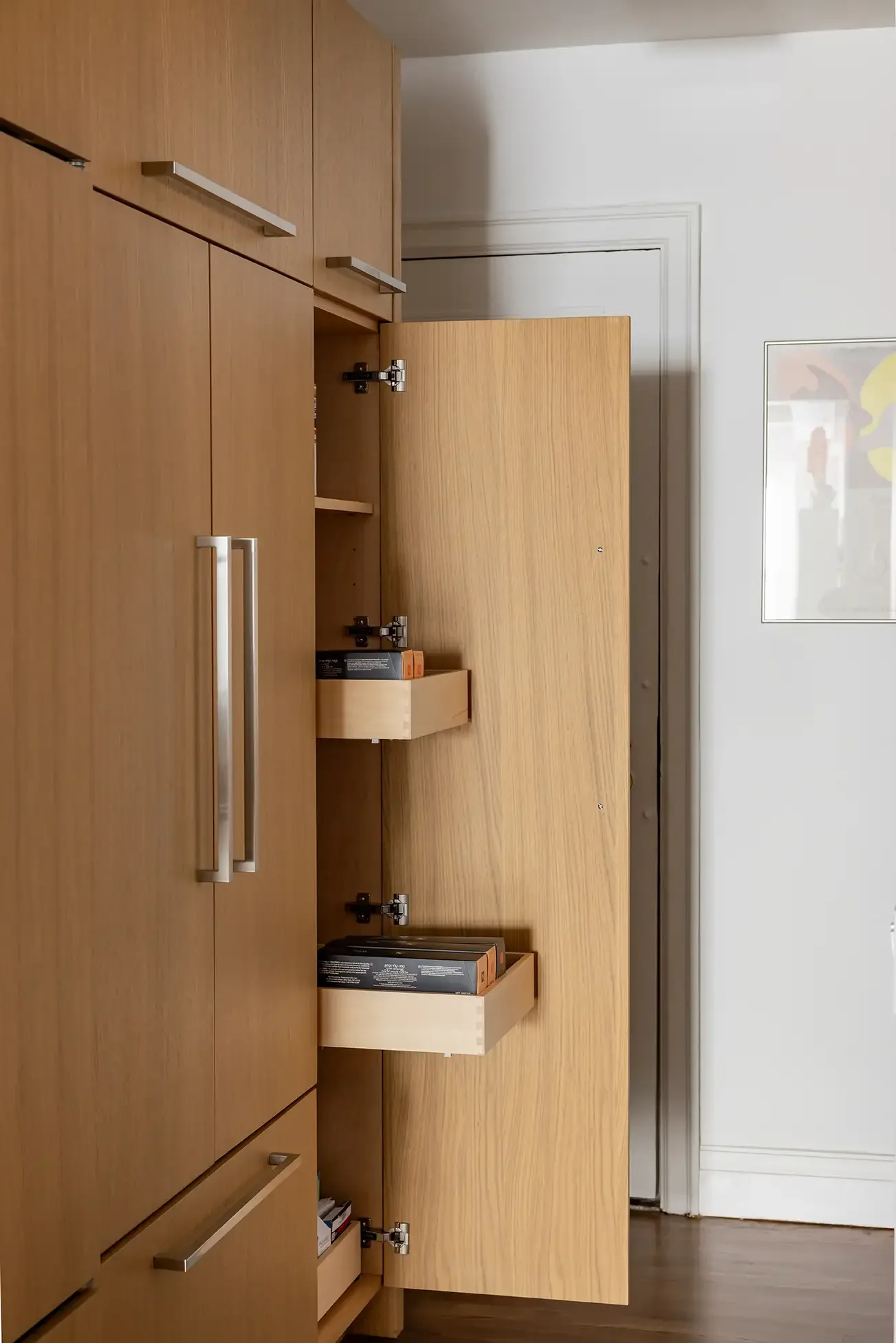 Open kitchen cabinet with pull-out drawers containing canned and boxed food, made of light wood with metallic handles.