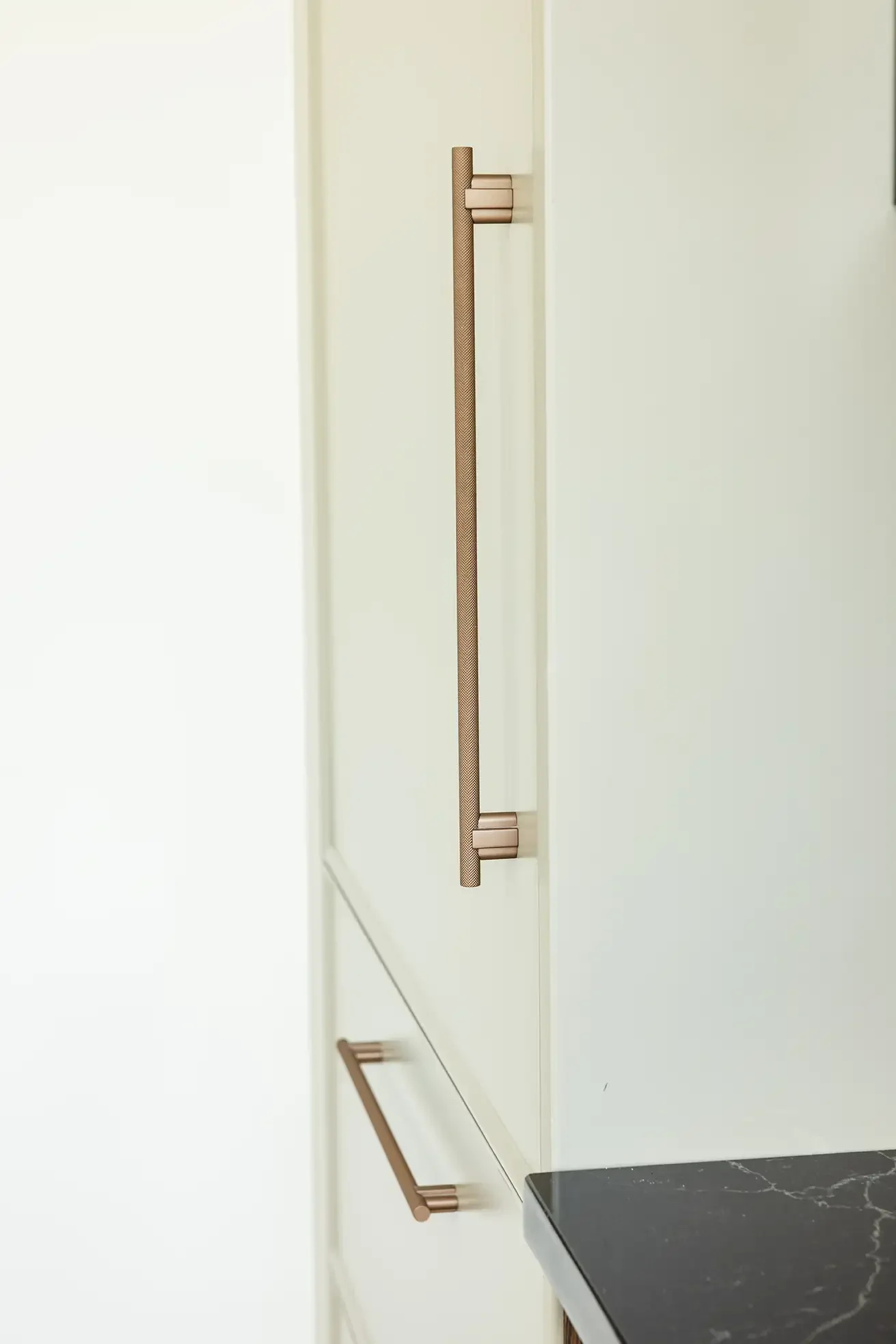 Close-up of gold-colored cabinet handles mounted on a white cabinet door and drawer, with a black marble countertop at the bottom right.