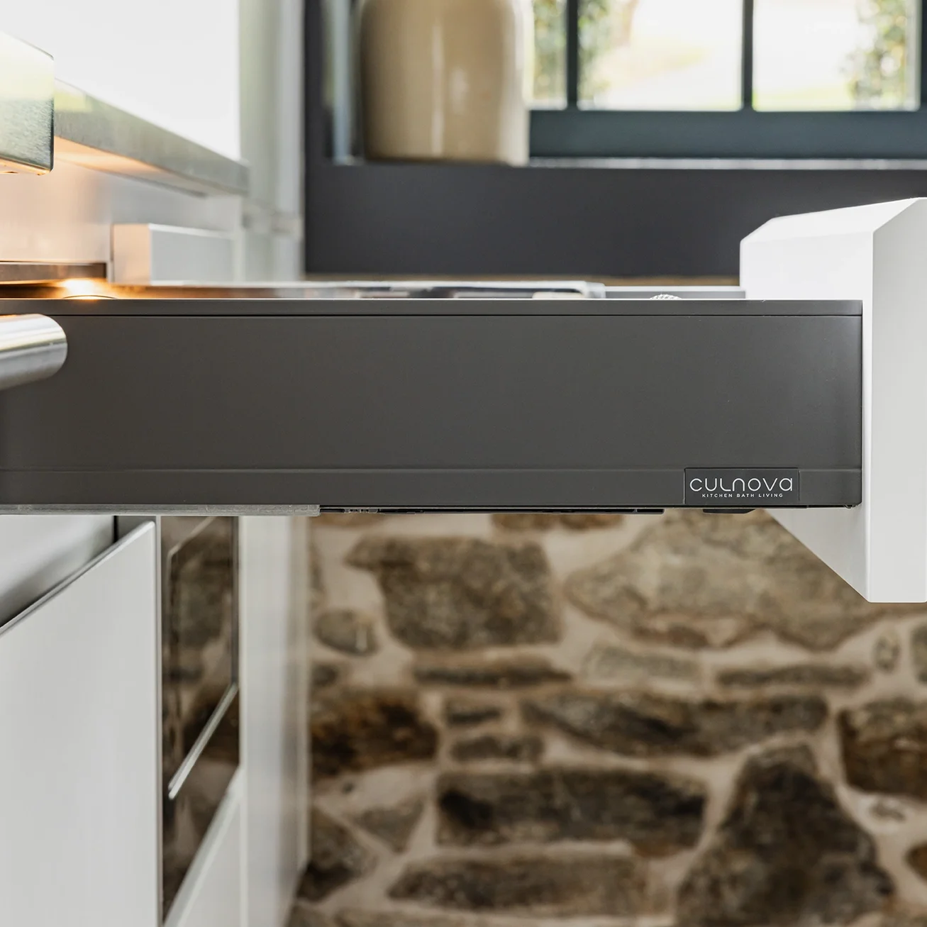 Close-up of a modern built-in oven or microwave with a dark gray finish and a small logo on the right side, installed in a white kitchen cabinet with a stone wall background.