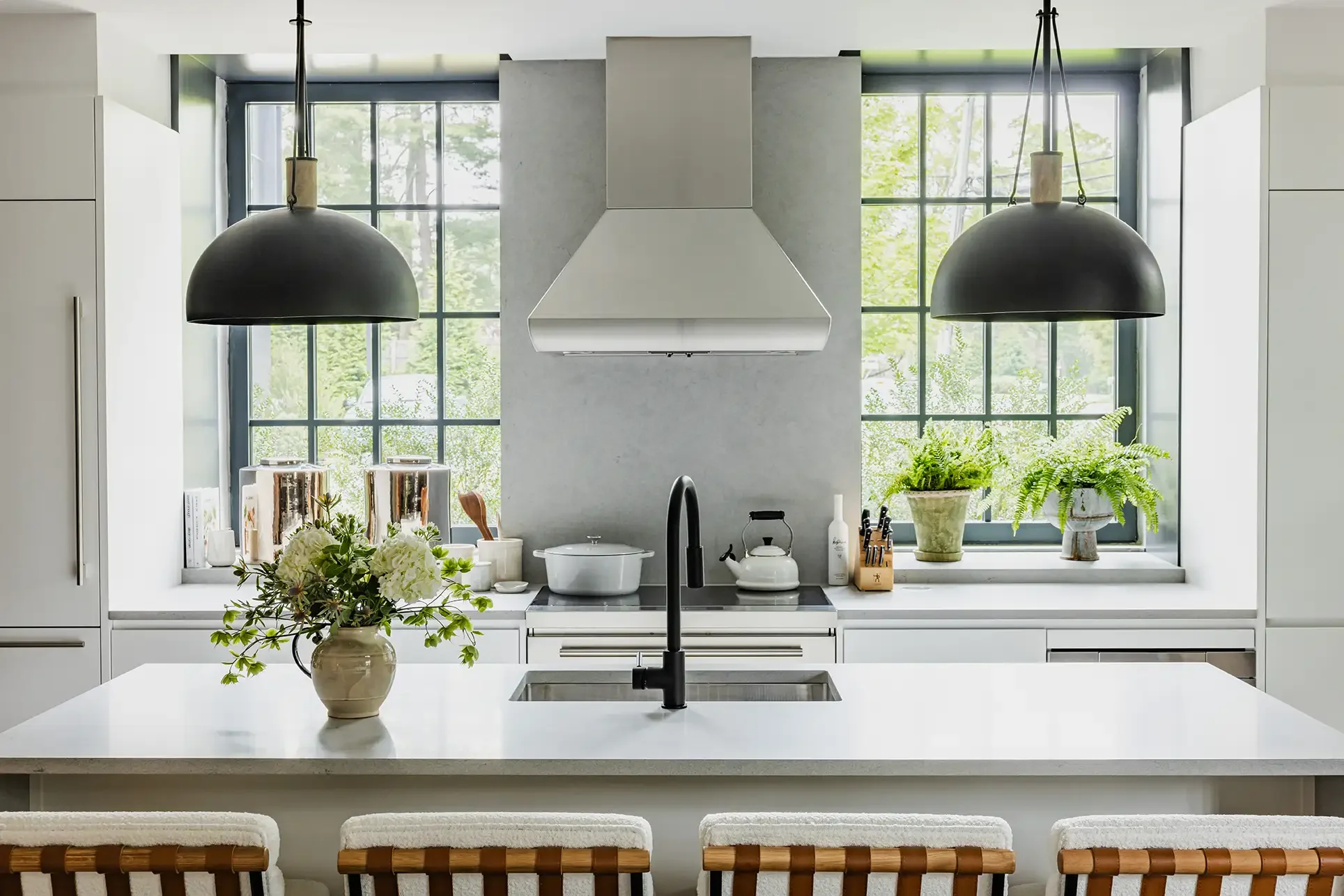 Modern kitchen with large black pendant lights, white cabinetry, a central island with a flower vase, a black faucet, green plants by the window, and cooking utensils.