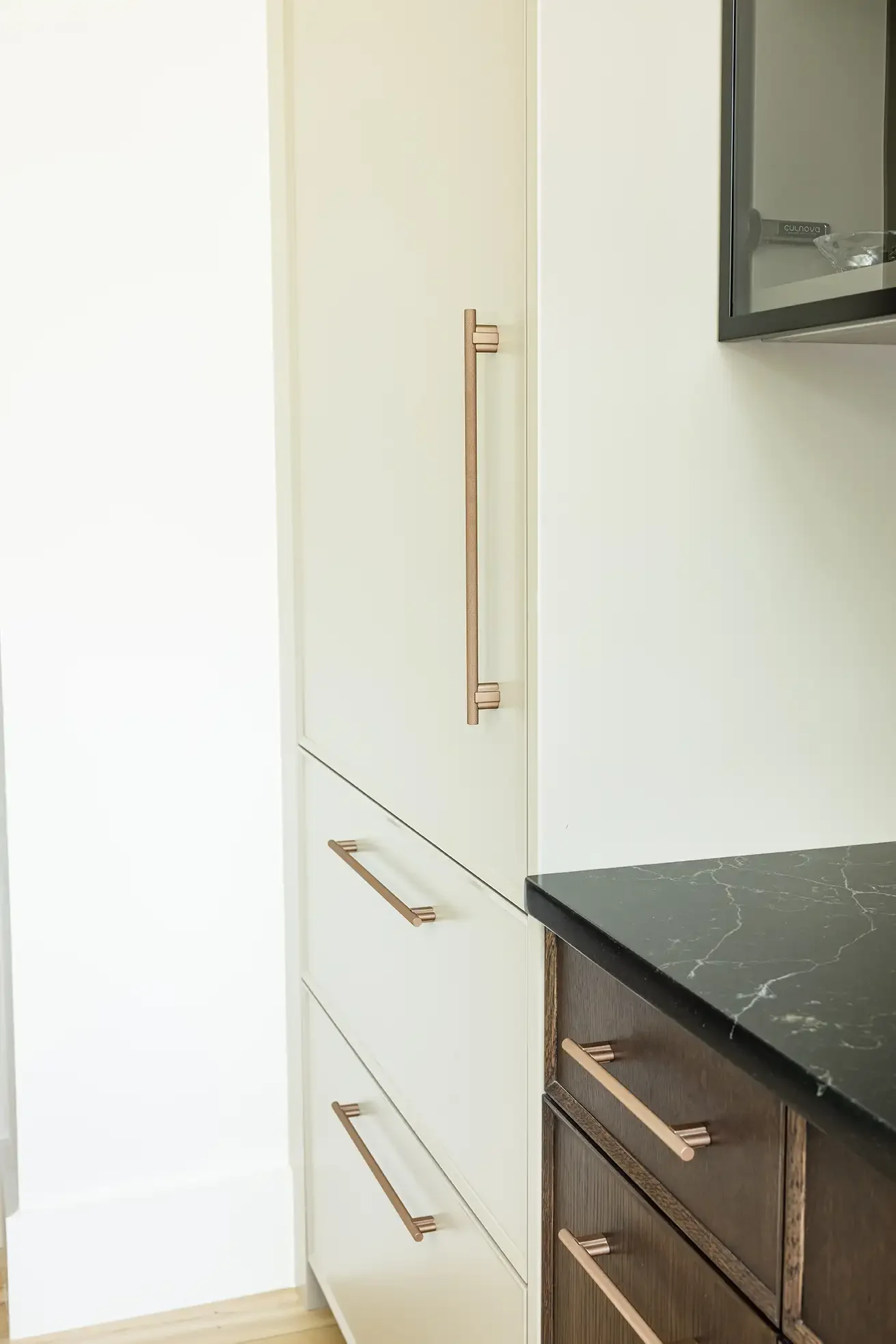 Close-up of a kitchen cabinet with vertical and horizontal copper-colored handles, next to a granite countertop.