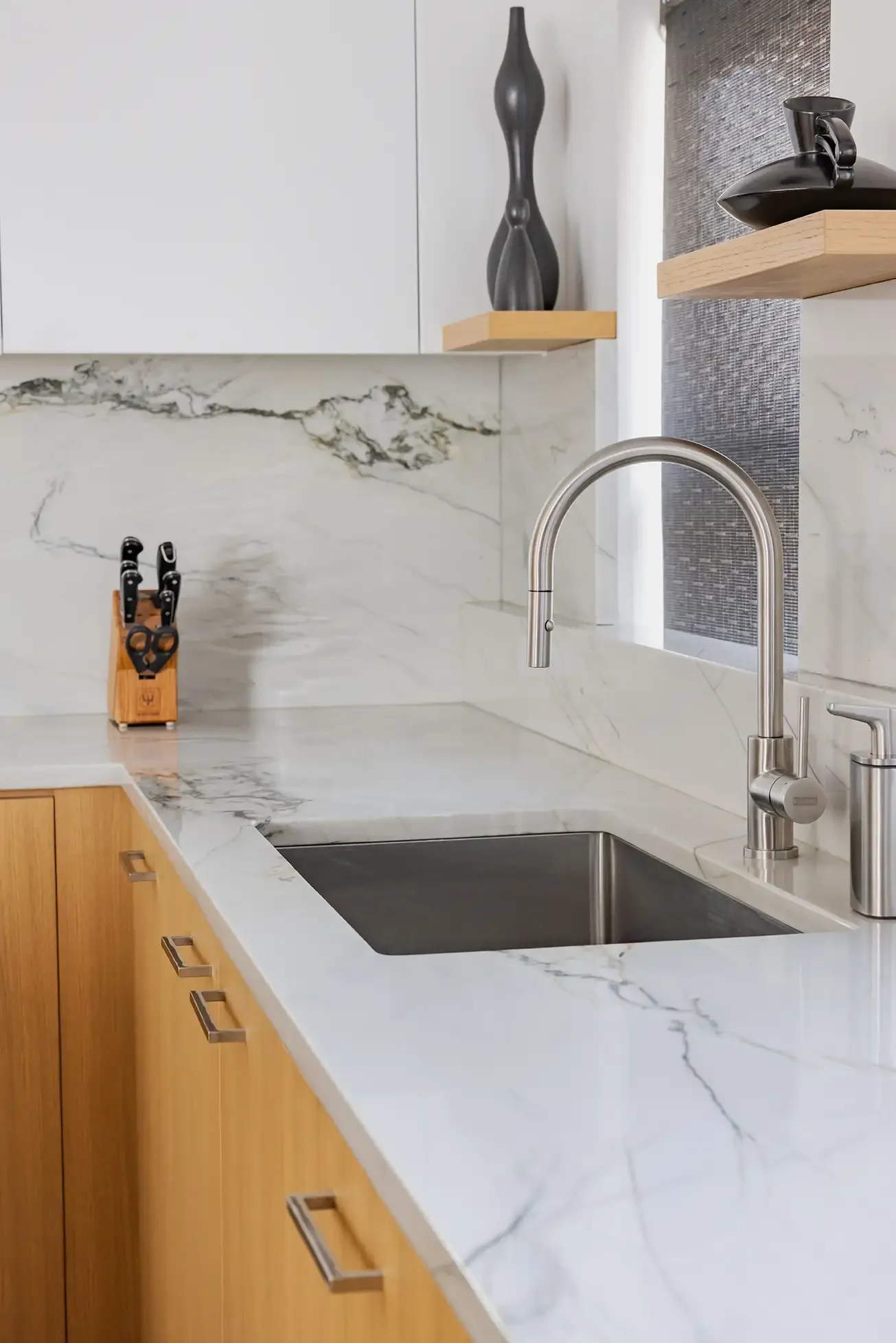 A modern kitchen with white marble countertops and backsplash, wooden cabinets, a stainless steel faucet, a knife block, and decorative vases on wooden shelves.