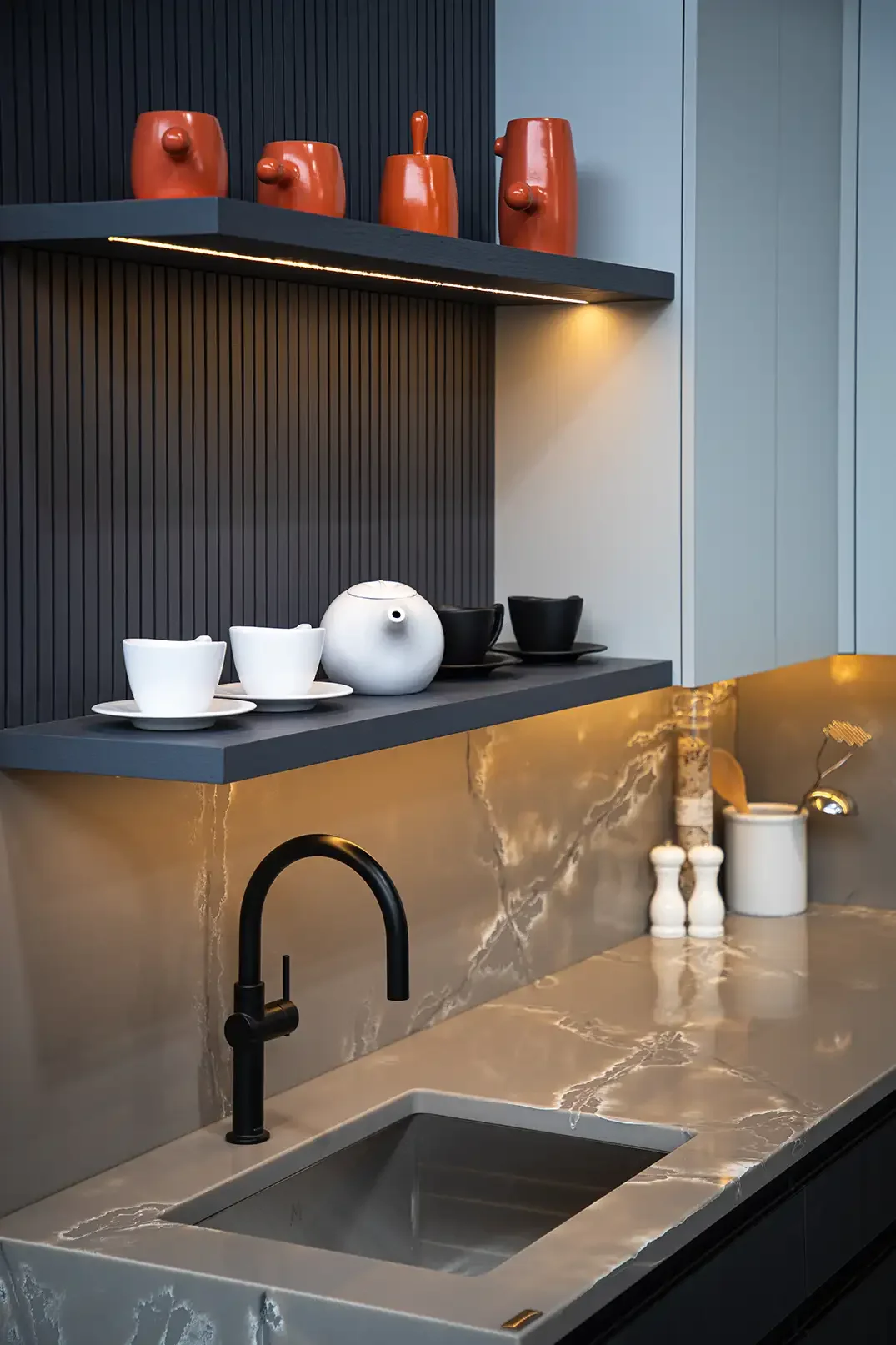 Modern kitchen backsplash with black textured wall, gray cabinets, black faucet, and marble countertop. Decorative red vases on upper shelf, white and black teapots on middle shelf, and white pepper and salt shakers and a utensil holder on the counte