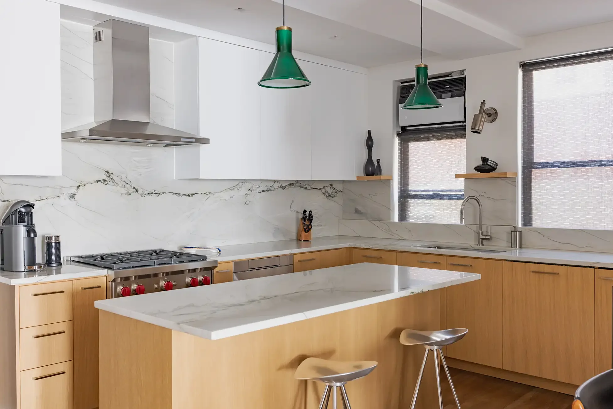 Modern kitchen with white upper cabinets, wooden lower cabinets, a marble backsplash and countertop, two green pendant lights, stainless steel appliances including a gas stove and coffee machine, and a kitchen island with marble top and two wooden st