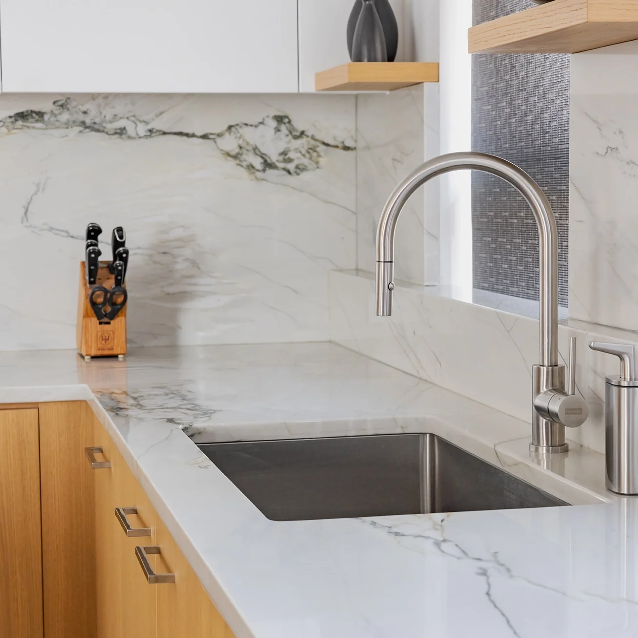 Modern kitchen countertop with a stainless steel faucet and a black sink, wooden cabinets, a knife block with knives, and marble backsplash with white and gray veining.