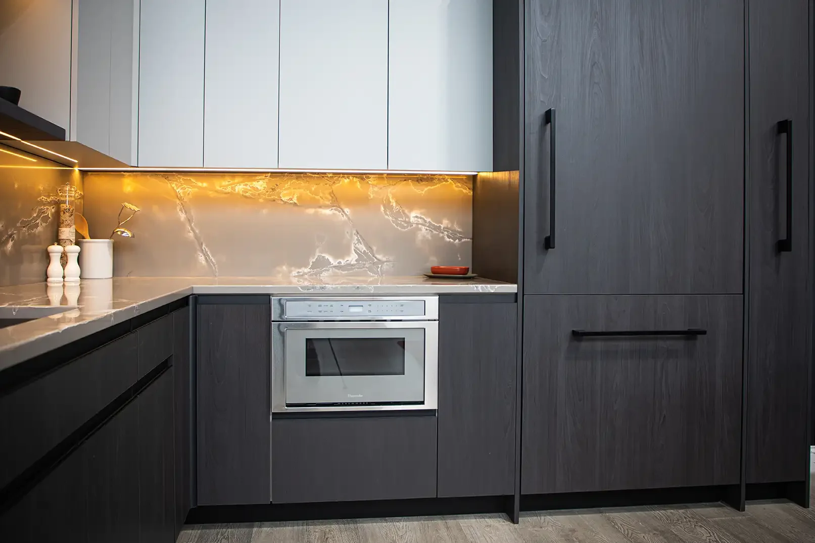 Modern kitchen with dark wood cabinets, marble countertops, and a built-in oven, illuminated by under-cabinet lighting.