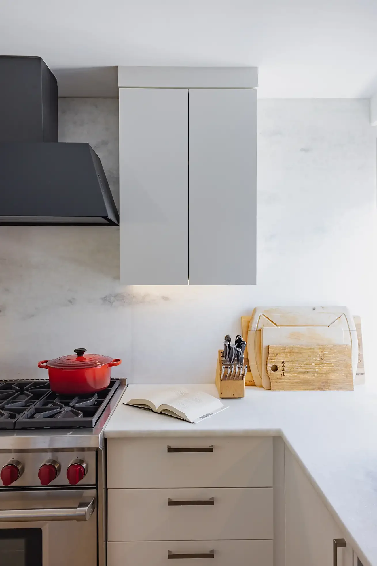 Modern kitchen countertop with a red pot on a stove, open book, knife set in a wooden block, and cutting boards against a white wall with gray cabinets.