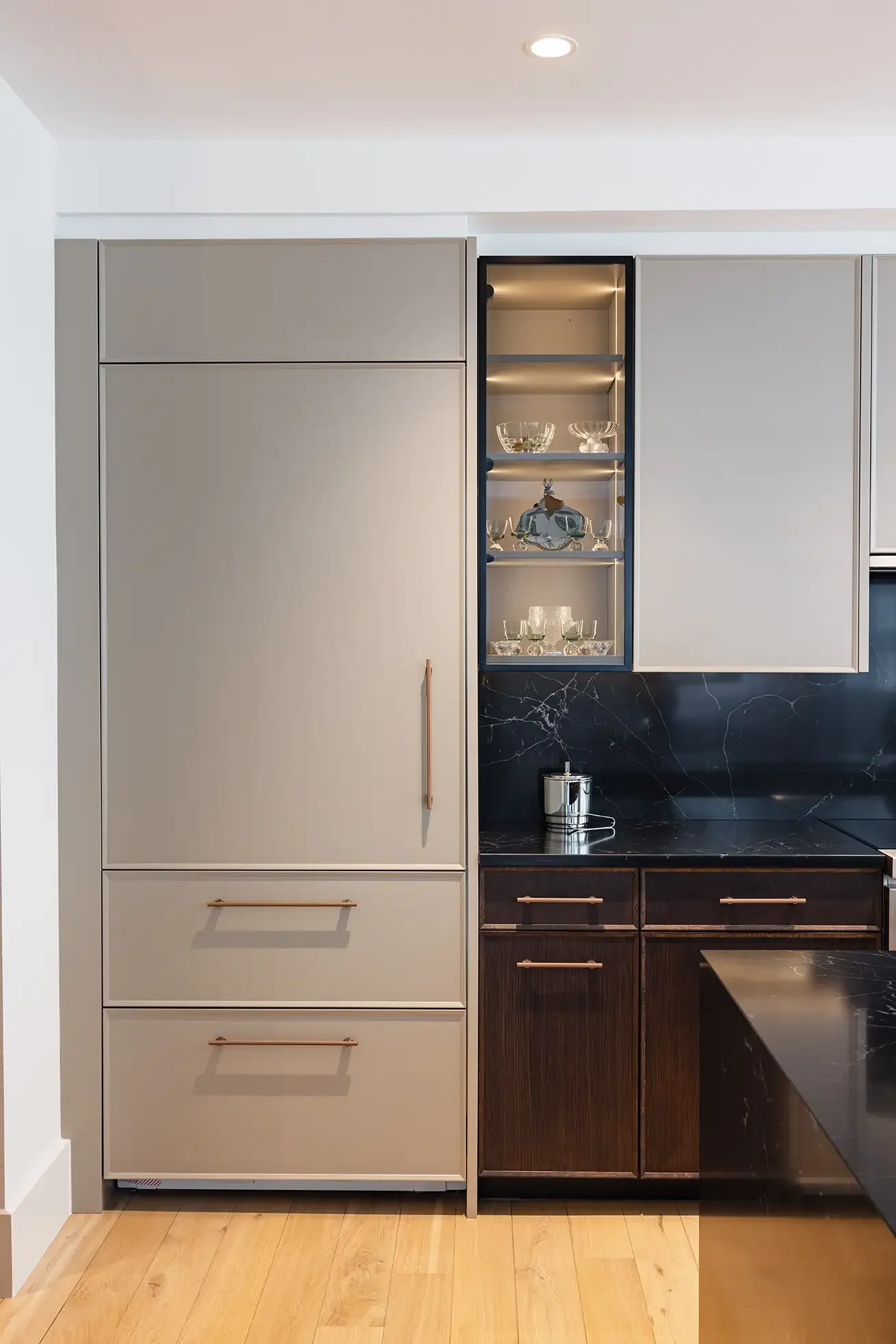 Modern kitchen with beige and dark wood cabinets, black marble countertop and backsplash, glass display shelves with glassware, and light wood floor.