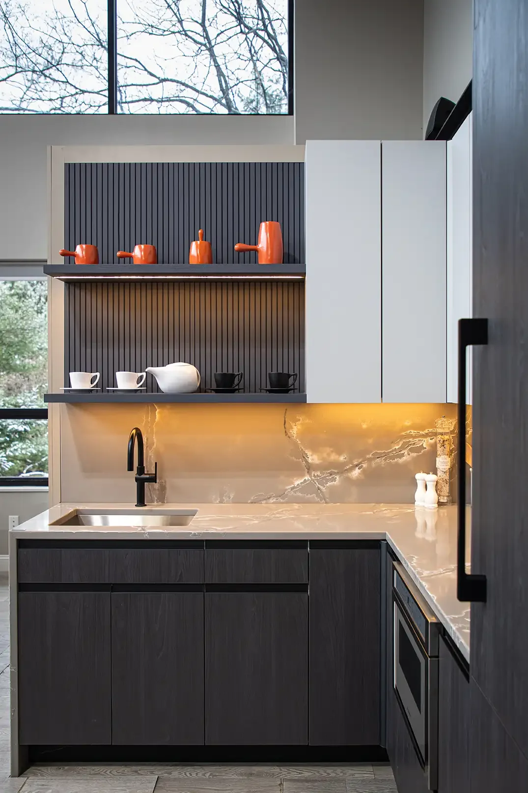 Modern kitchen with black lower cabinets, white upper cabinets, marble backsplash, open shelves with orange and black teapots and cups, black faucet, and large window showing tree branches outside.
