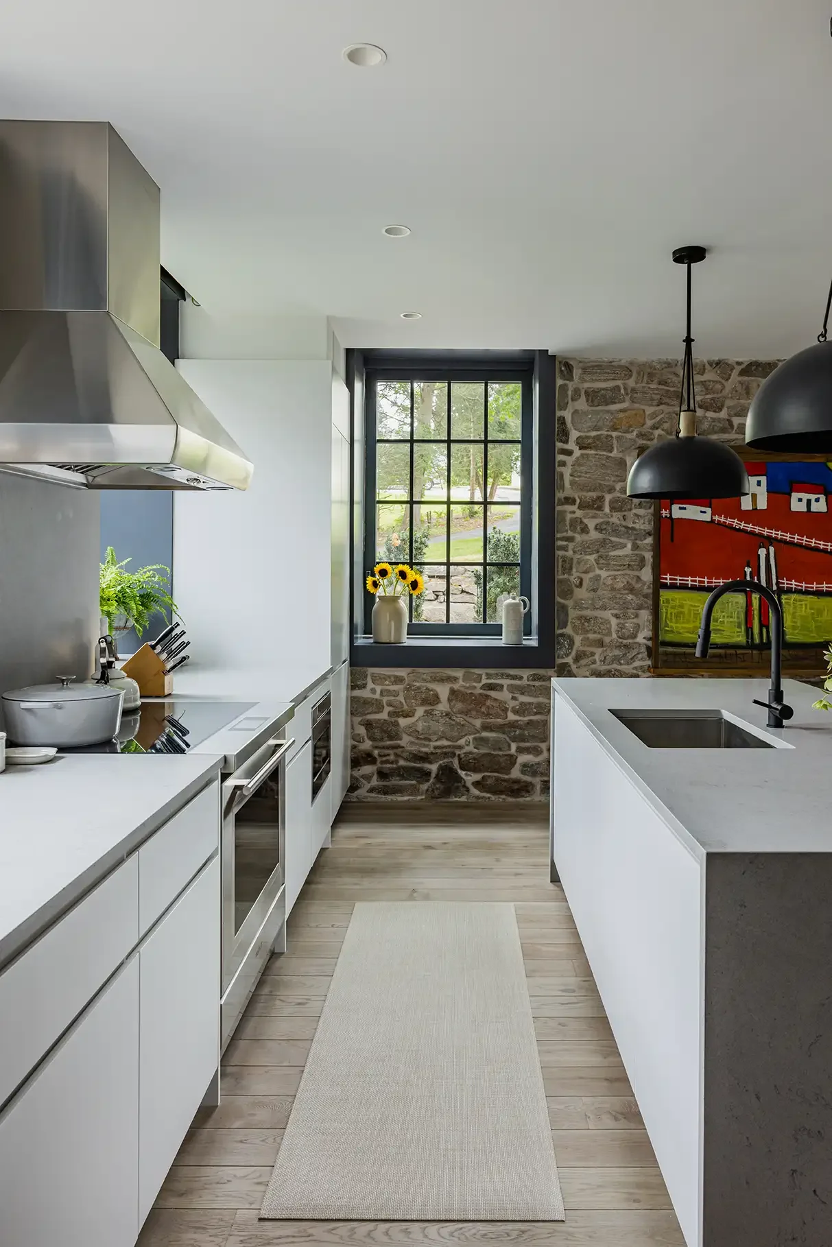 Modern kitchen with white cabinets, stainless steel appliances, a black faucet, black pendant lights, a window with a stone wall, and a colorful painting on the wall.