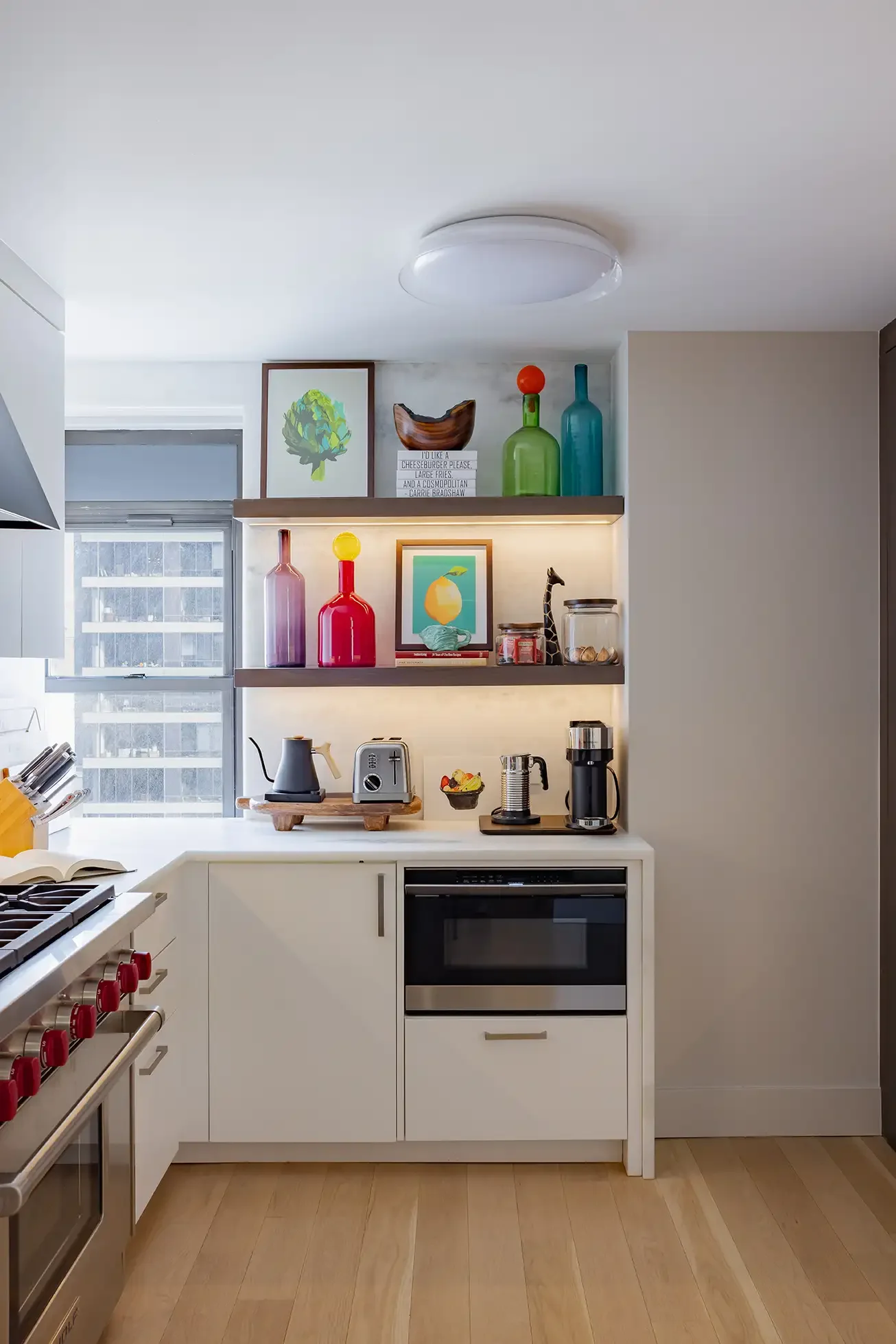 Modern kitchen with white cabinetry, wooden shelves displaying colorful glass bottles, framed artwork, and kitchen appliances like a toaster, electric kettle, and coffee maker. There is a window with blinds and a built-in oven.