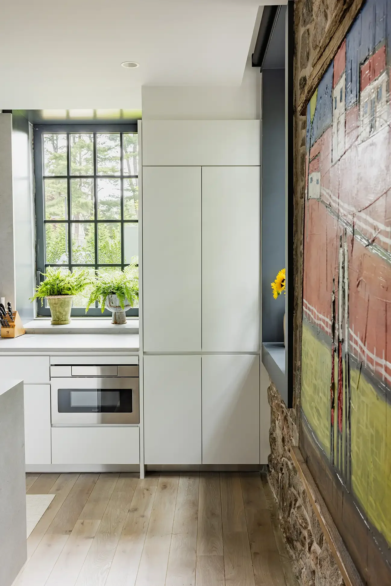 Modern kitchen with white cabinets, a window with grass outside, potted plants, a built-in microwave, a colorful abstract painting on the wall, and light wood flooring.
