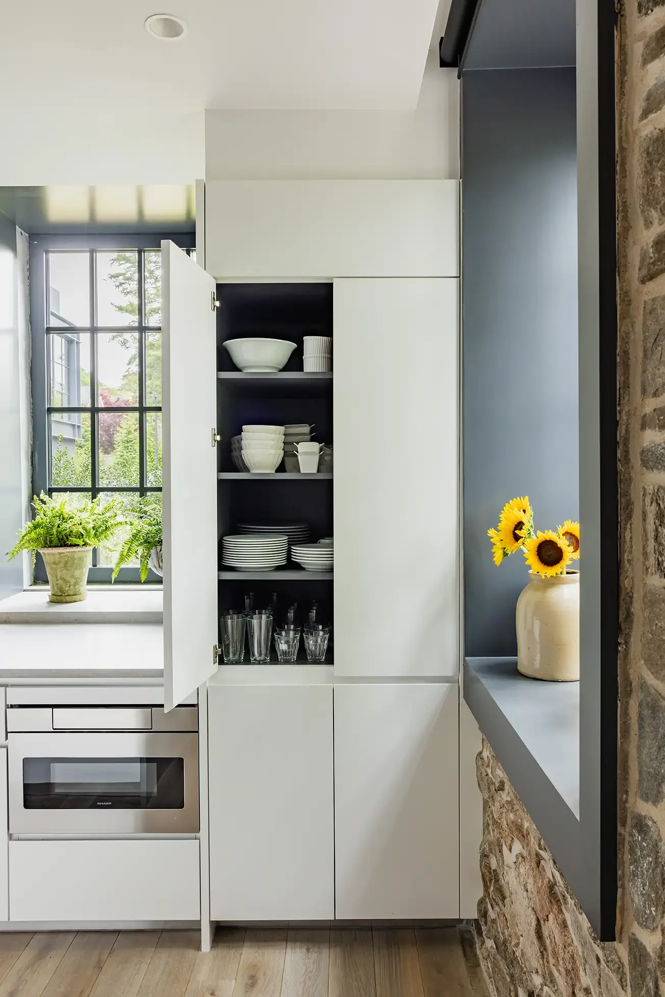 Open kitchen cabinet filled with white dishes, glasses, and bowls, next to a window with a potted fern and a jar of sunflowers.