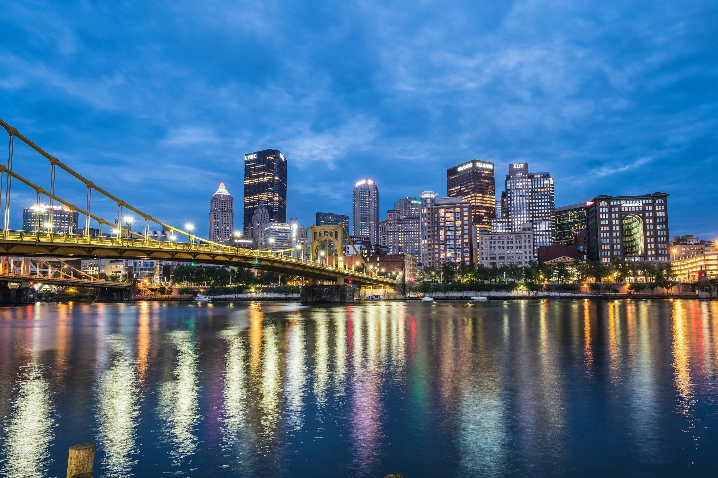 Nighttime Pitsburgh city skyline with tall buildings, a bridge, and reflections on the water.